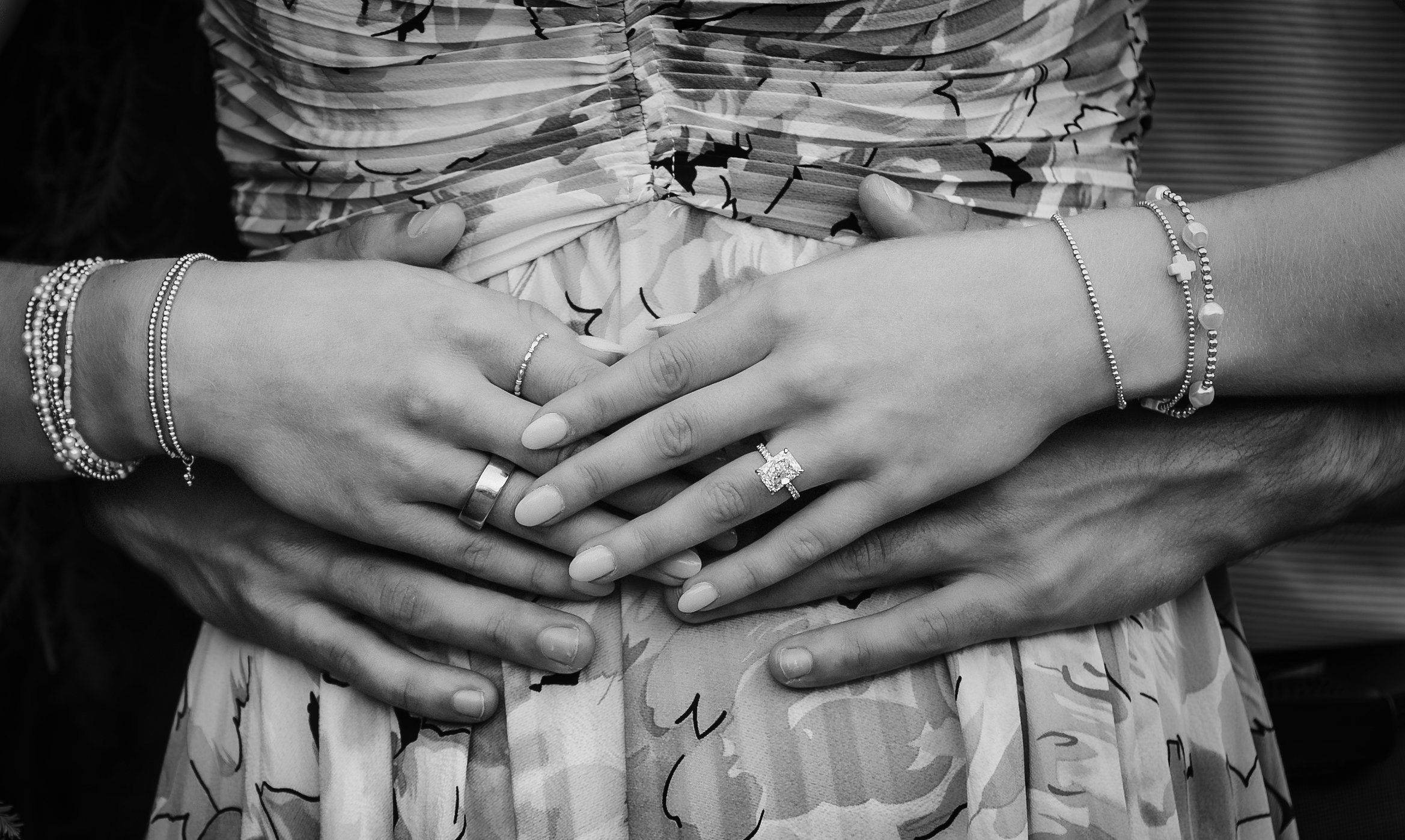 Two pairs of hands, one with a ring and several bracelets, resting on the stomach of a person wearing a floral-patterned dress. The other person has their hands over the first person's hands, also wearing bracelets.
