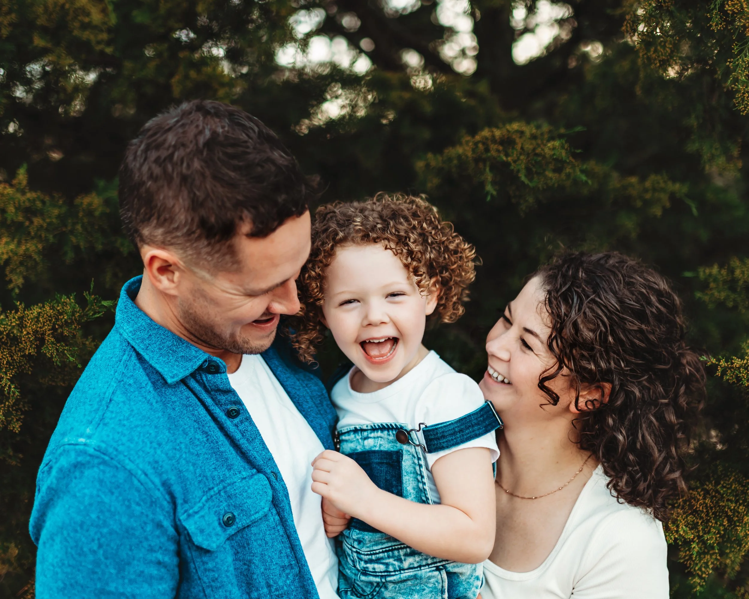 A happy family of three, consisting of a father, mother, and young child, enjoying time together outdoors. The father is smiling while looking down at the child, who is laughing, and the mother is smiling at the child as well. They are standing close
