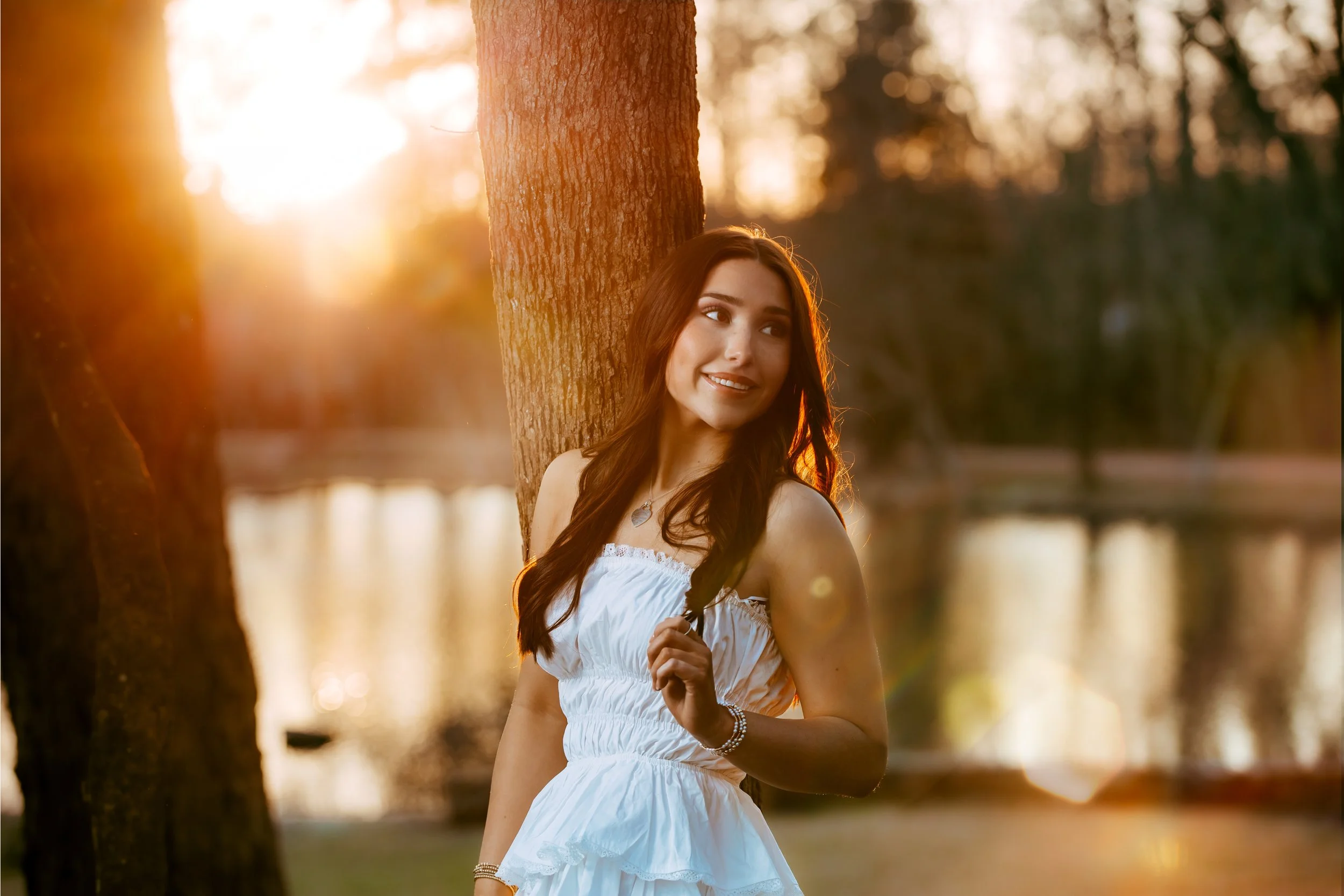 A young woman with long dark hair is standing outdoors near a tree, smiling and looking to the side during sunset, with a river in the background.