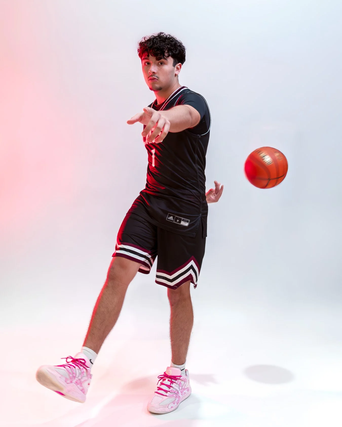 Young man in black basketball jersey and shorts, wearing pink and white sneakers, is playing basketball on a white background, with a red and orange basketball in mid-air.