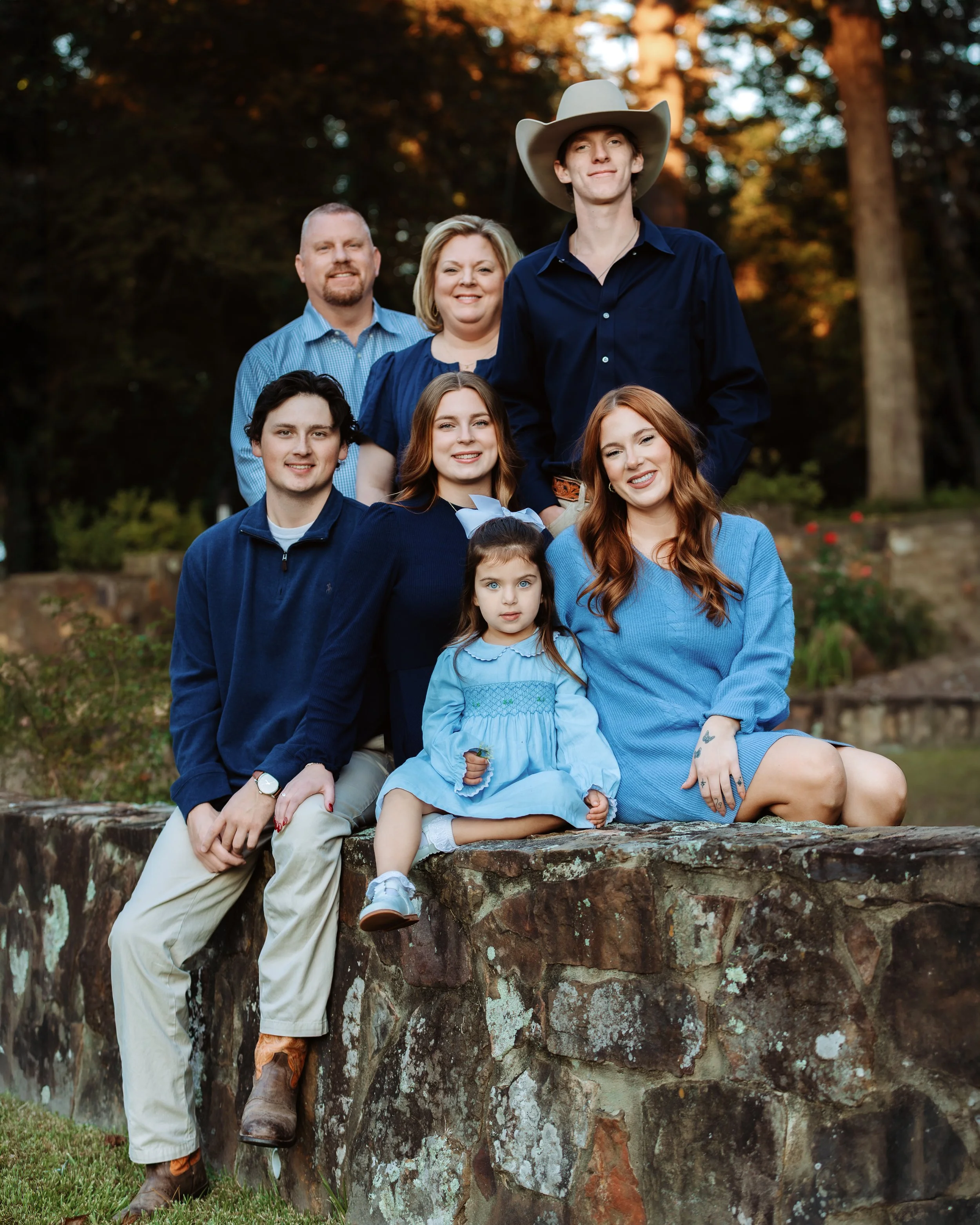Family photo of seven members outdoors on a stone wall, dressed in blue and neutral tones, with trees and sunset in the background.