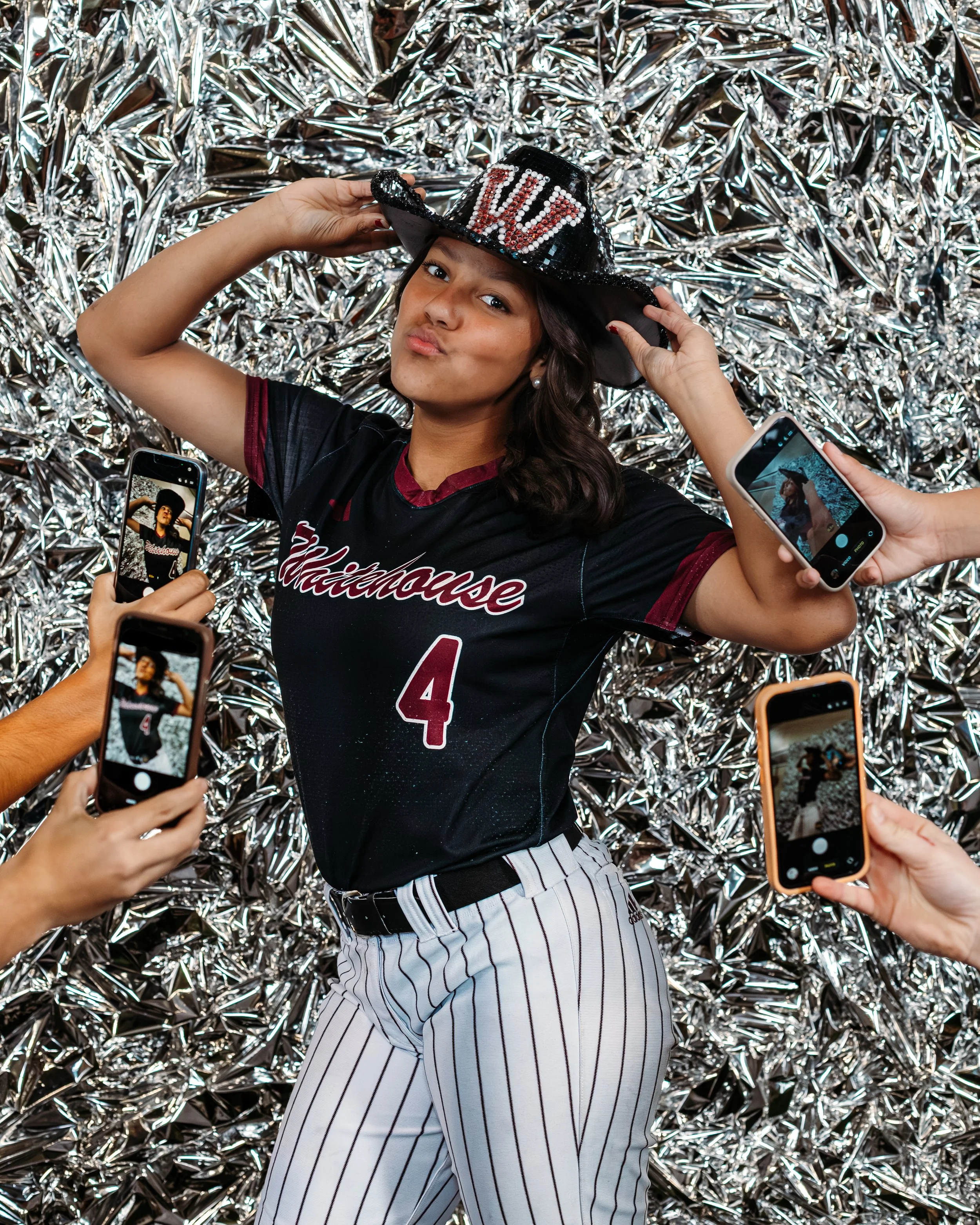 A woman in a black and red baseball jersey with the number 4, striped white and black pants, and a shiny black hat with the word 'LOVE' in sequins, posing with a puckered lips expression against a silver, metallic, crumpled background while being pho