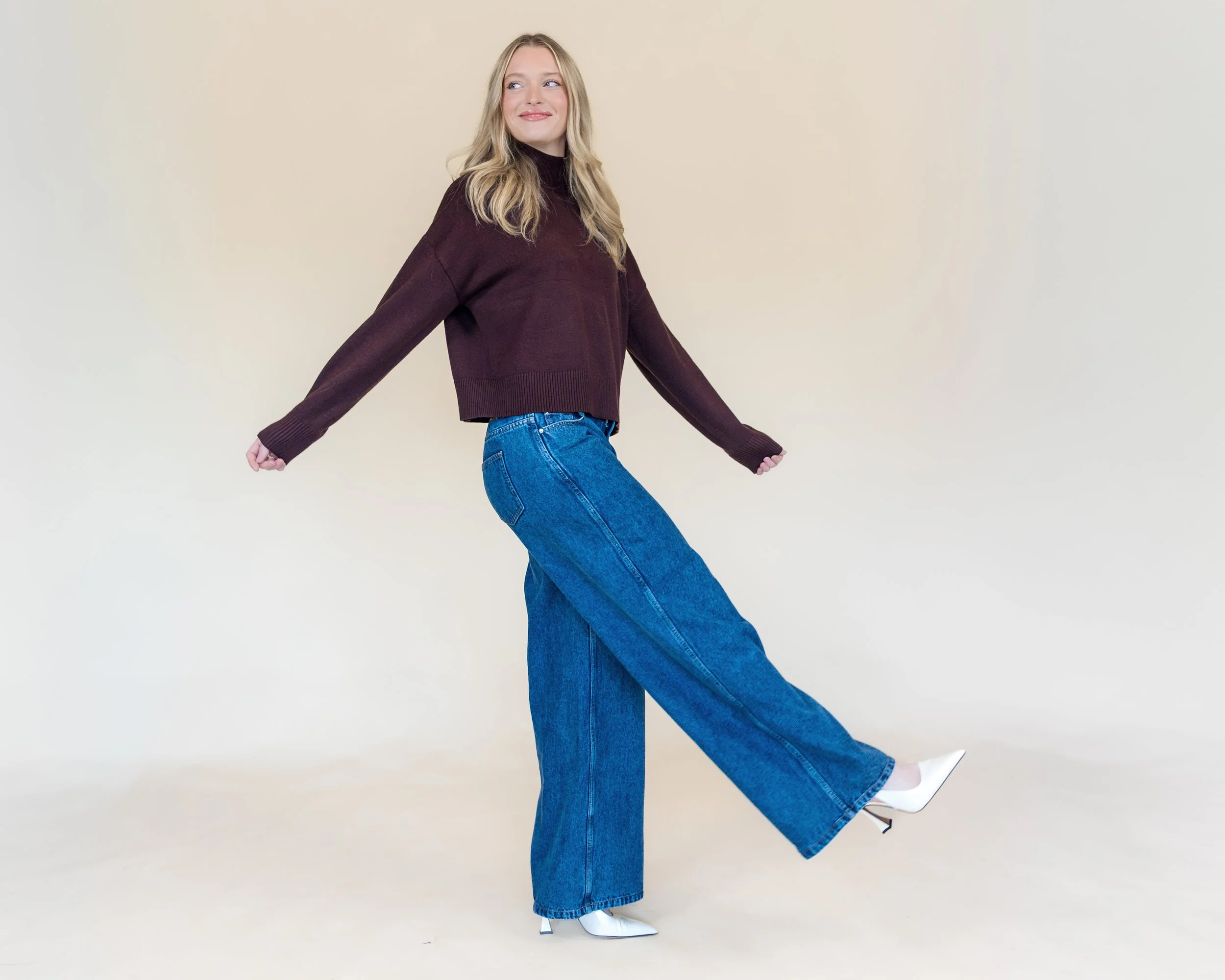 A woman with blonde hair posing in a casual outfit against a neutral background, wearing a burgundy sweater, blue wide-leg jeans, and white high-heeled shoes.