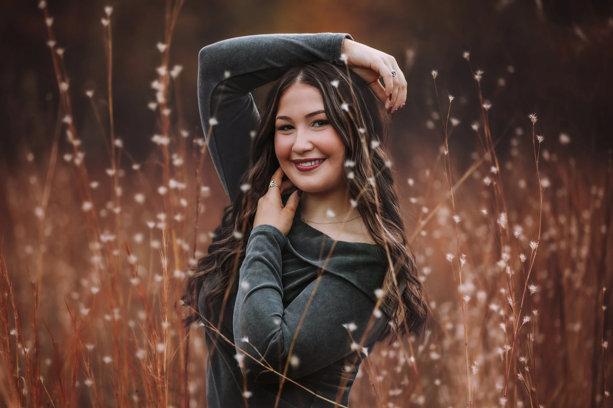 A young woman with long brown hair smiling, standing in a field of tall, dry grass or plants with white fluffy tops, during sunset or golden hour.