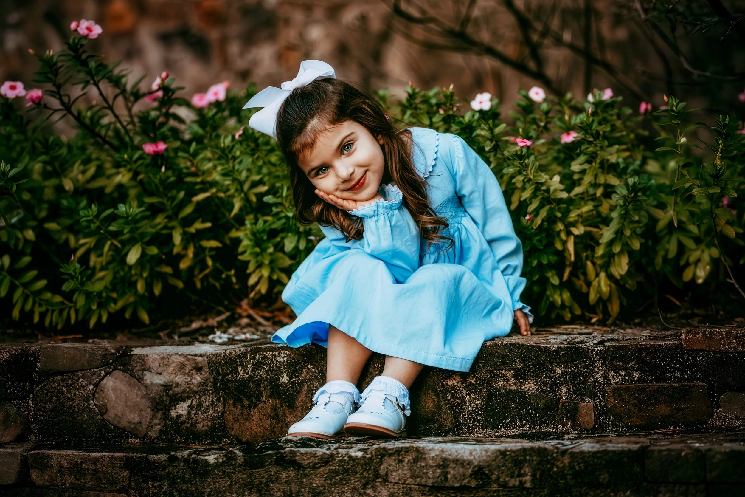 A young girl with brown hair, blue eyes, and a white bow in her hair, sitting on outdoor steps, leaning her cheek on her hand, smiling, surrounded by green bushes with pink and white flowers, wearing a light blue dress and white shoes.