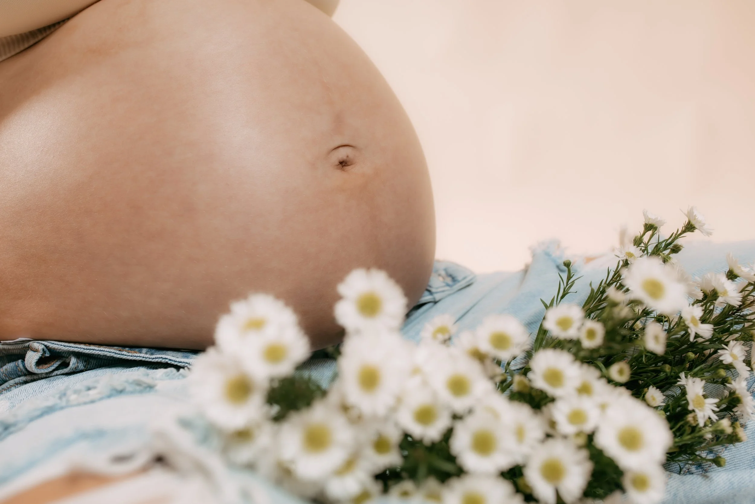 Maternity session of a pregnant belly with a small belly button, surrounded by a bunch of white daisies with yellow centers, resting on blue denim fabric.