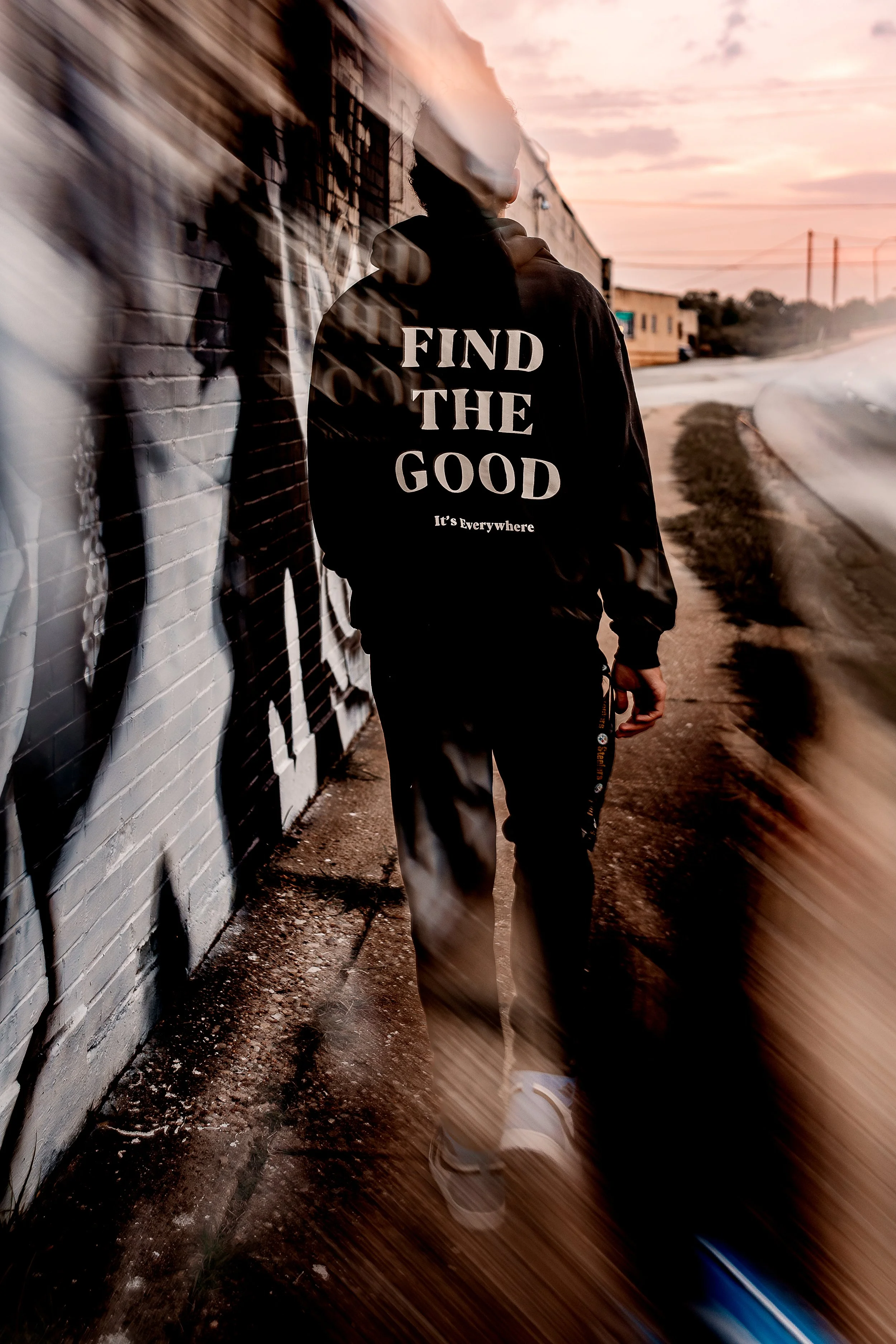 A person walking along a city sidewalk at sunset, wearing a black hoodie with the words 'FIND THE GOOD It's Everywhere' on the back, and dark pants. The person is facing away from the camera, passing by a graffiti-covered wall. The scene has an artis