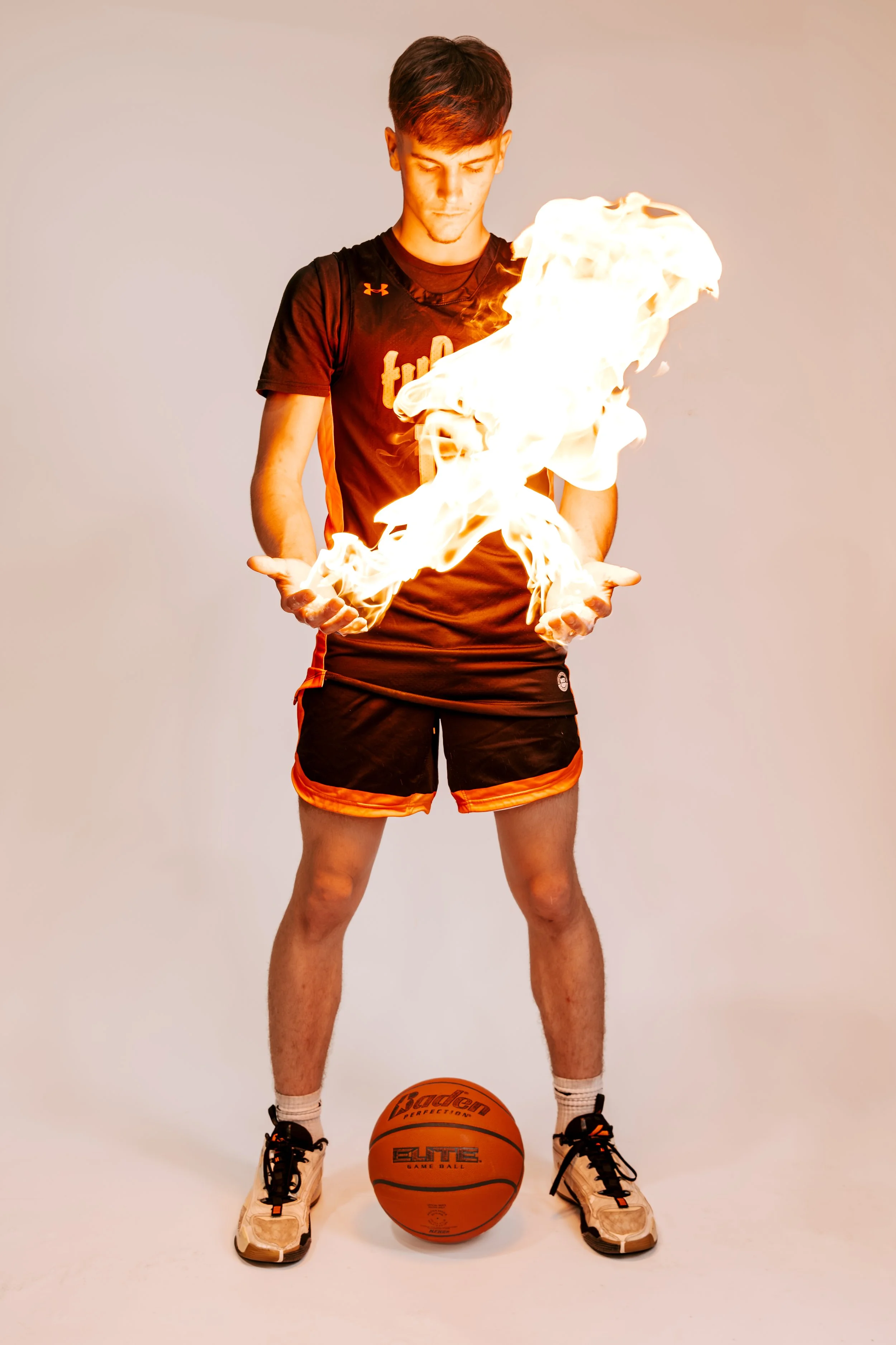 A young male athlete in a black and orange sports uniform is standing with his eyes closed, holding a flame in both hands, with a basketball on the ground in front of him, against a plain light background.