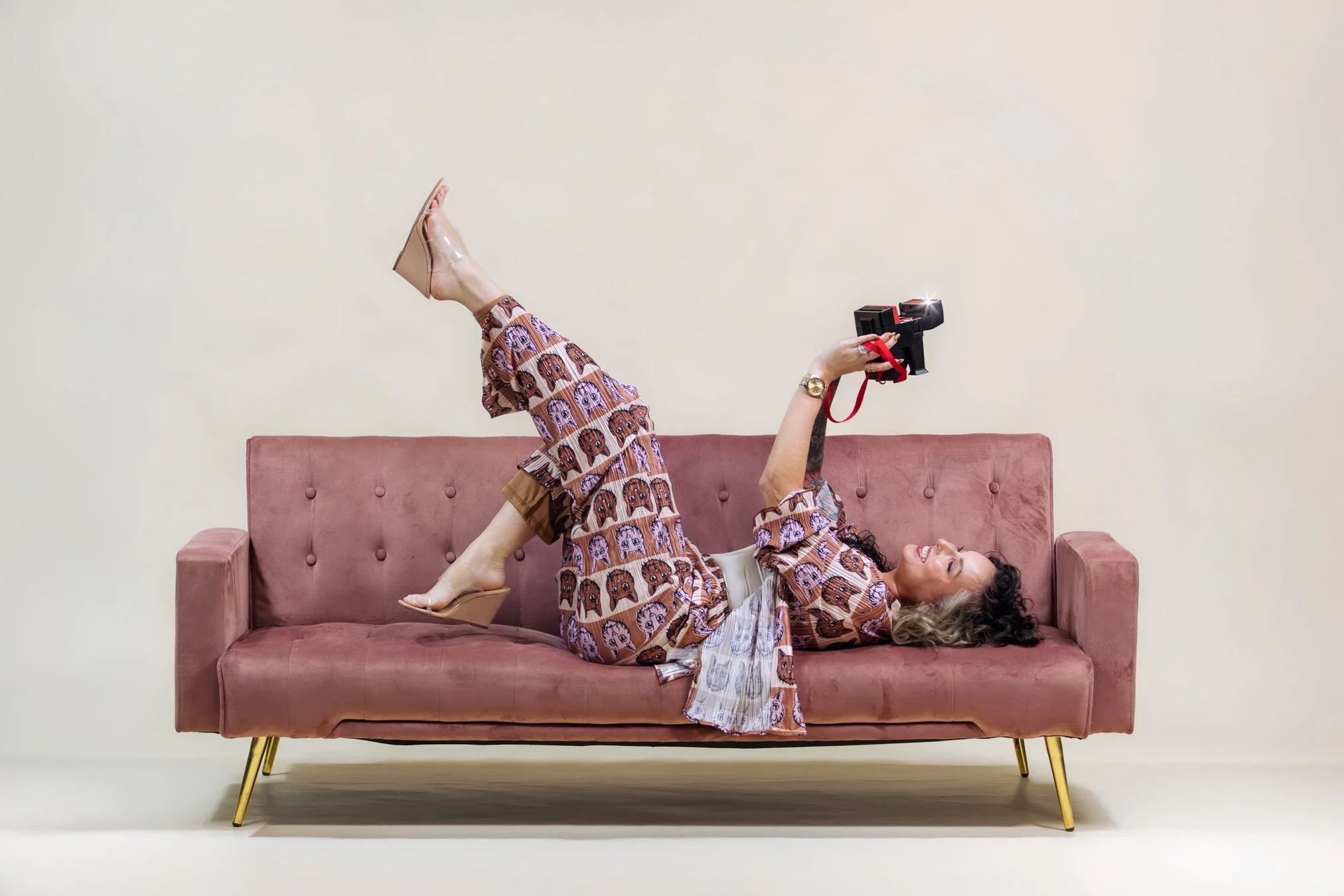 Woman lying on a pink sofa, wearing patterned pajamas, holding a camera, smiling, against a plain, light-colored background.