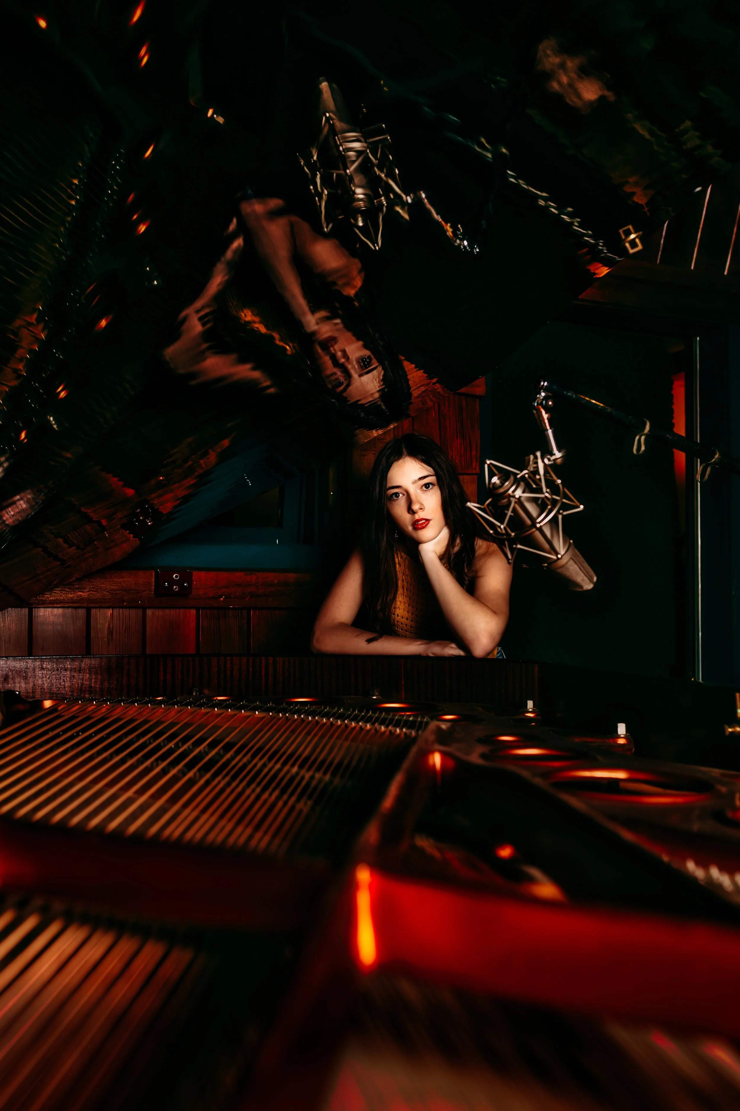 A woman with dark hair and red lipstick in a recording studio, leaning on a grand piano with a microphone nearby, illuminated by warm red and orange lighting.