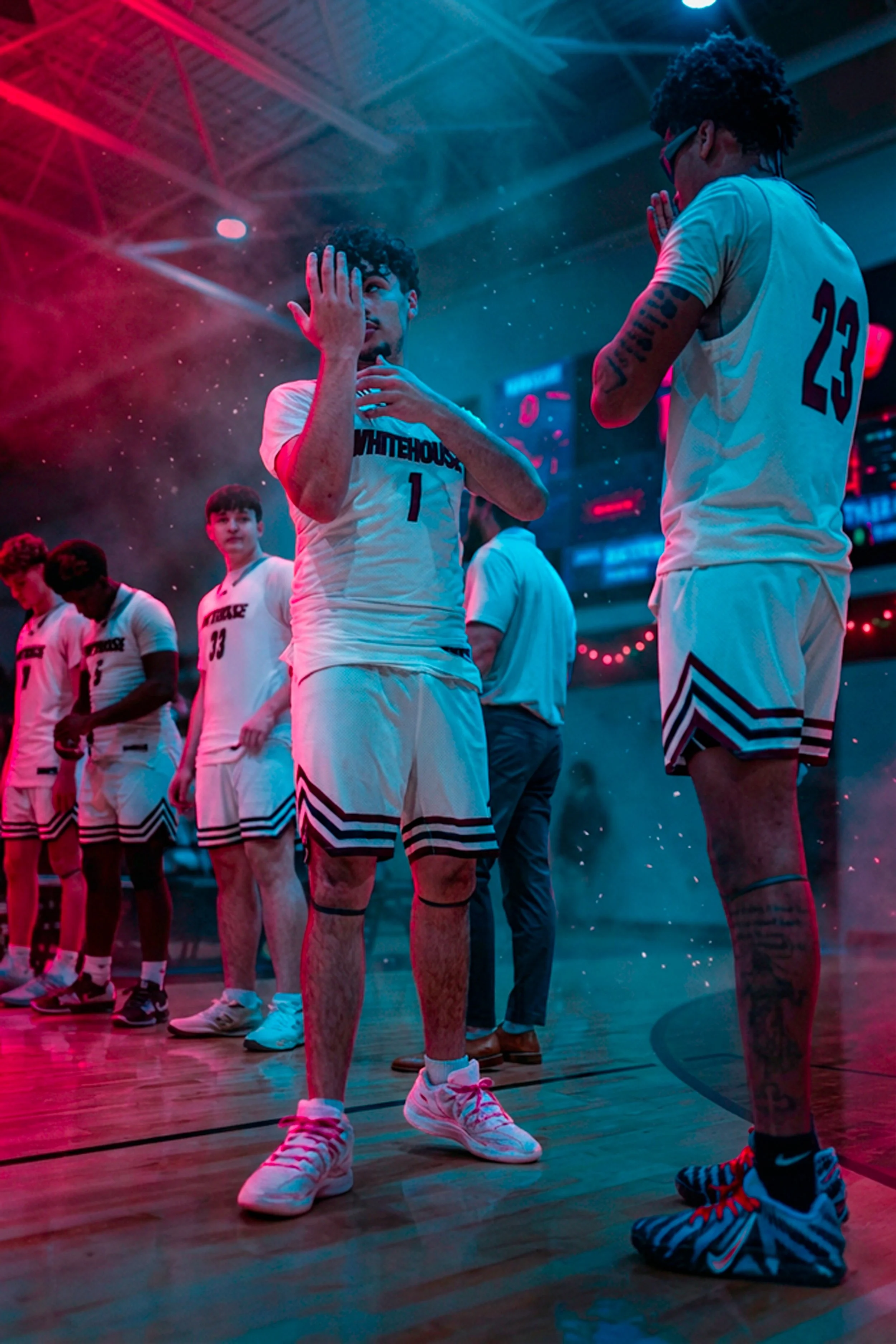 Basketball players in white uniform standing on a court with one player in the foreground adjusting his glasses and another player with tattooed arms praying or speaking softly, illuminated by colorful arena lights.