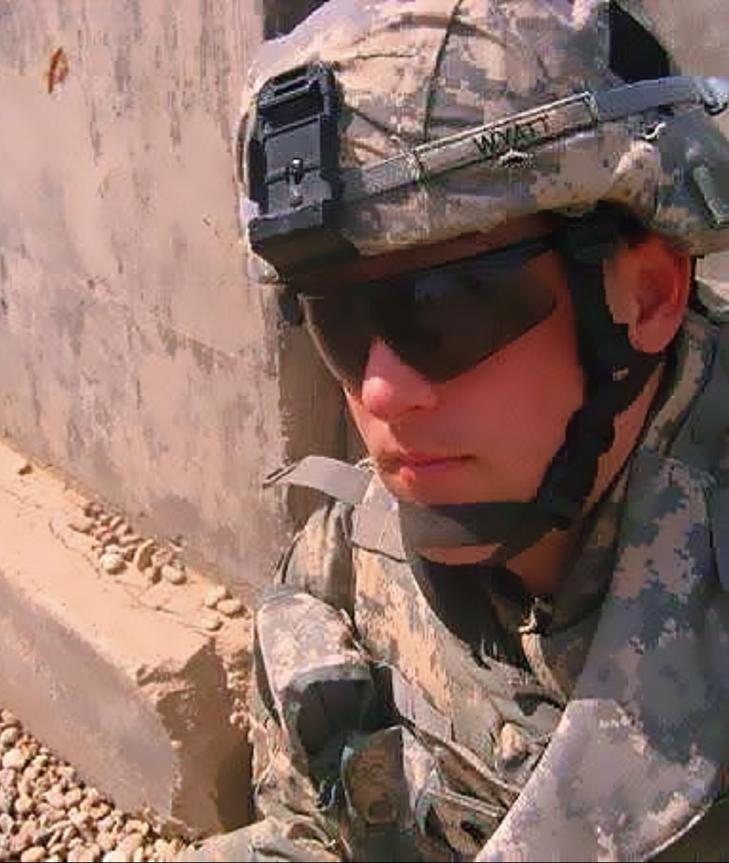 A soldier wearing desert camouflage uniform, tactical helmet, and sunglasses, sitting on the ground in a sandy environment.