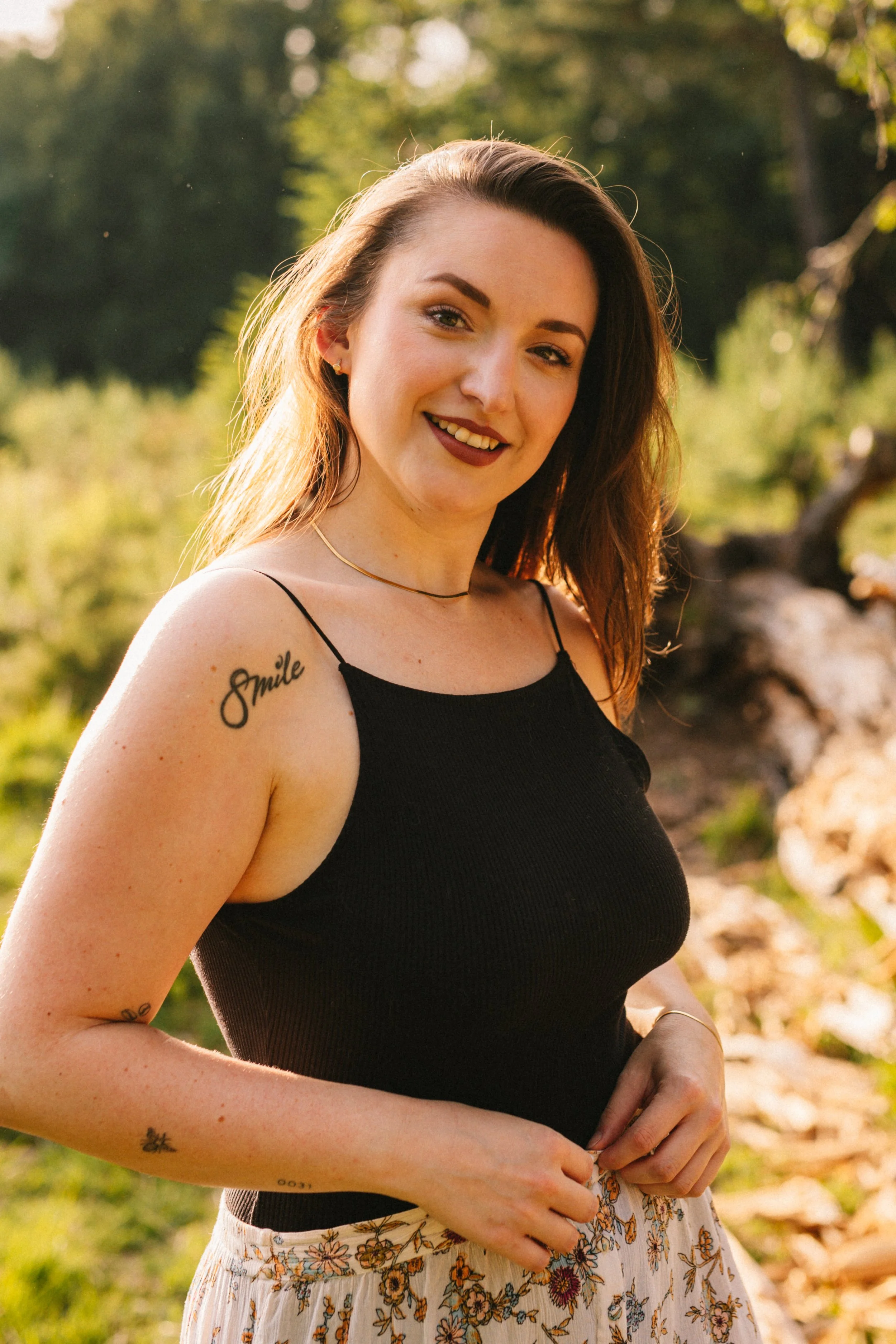 Eine lächelnde junge Frau im Sonnenlicht, trägt ein schwarzes Top und einen floralen Rock, steht im Freien in einer grünen, bewaldeten Umgebung.