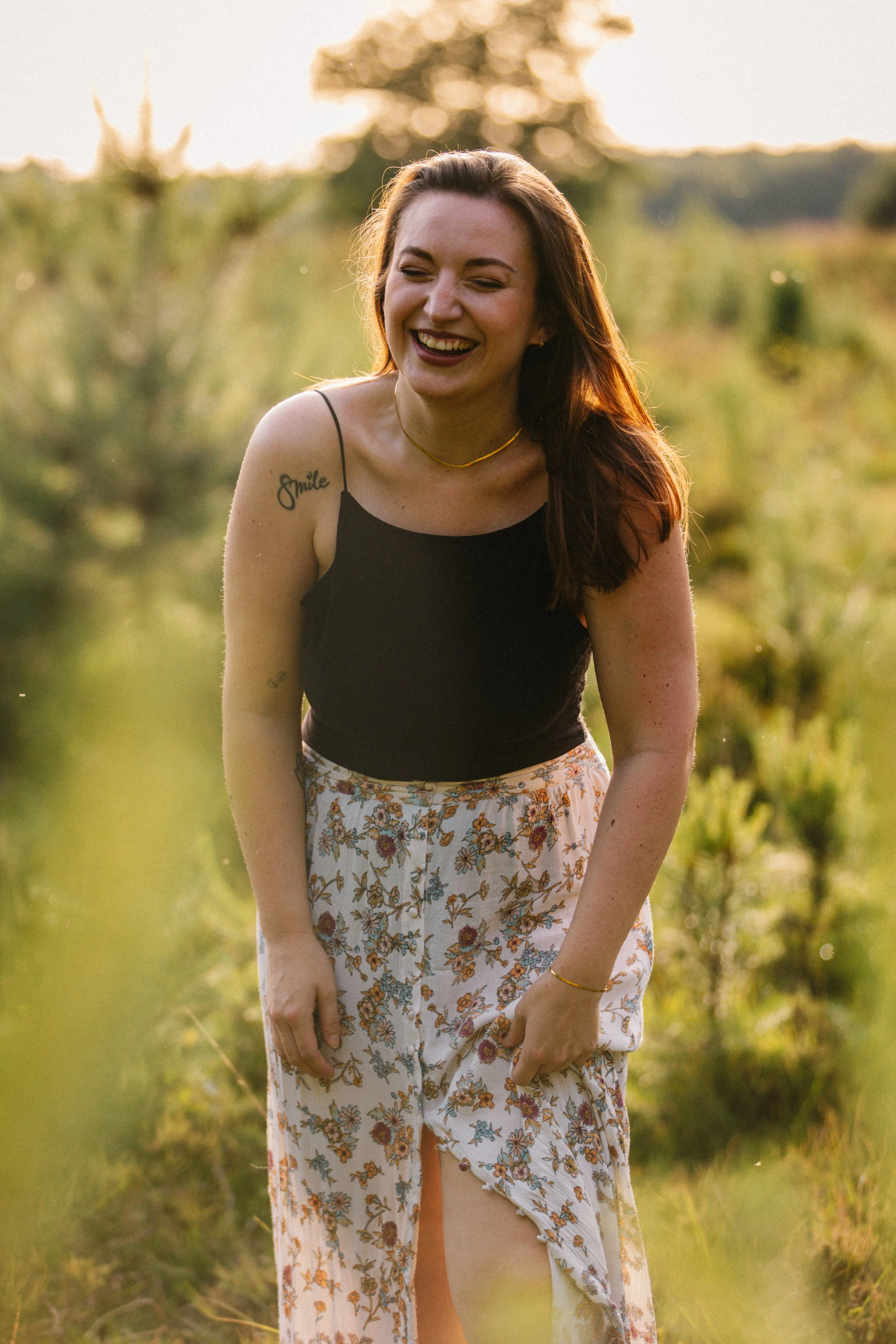 Eine lachende junge Frau in einem schwarzen Top und einer Blumenhose im Sonnenuntergang in der Natur.