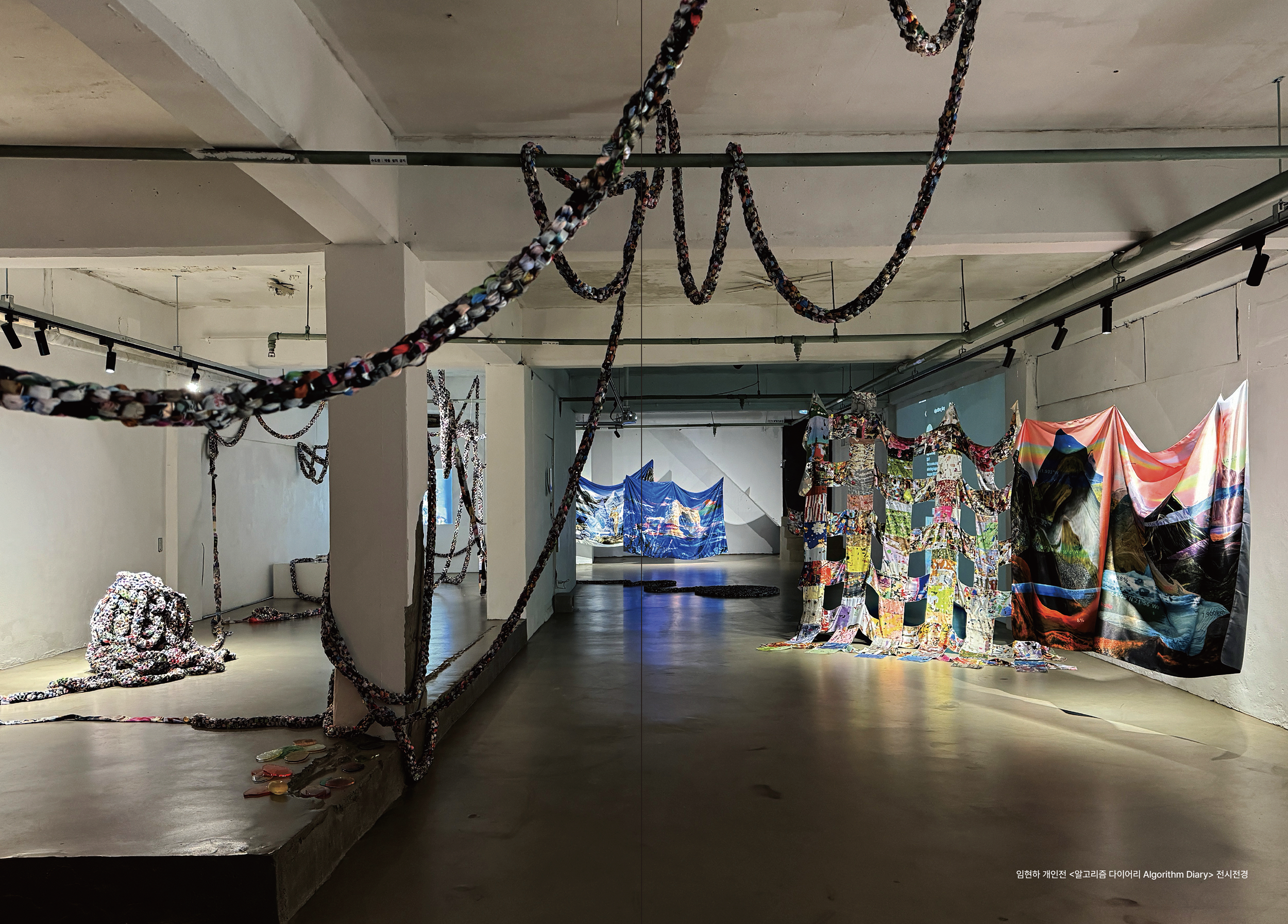 An art installation featuring colorful fabric art and braided ropes hanging from the ceiling in an industrial-style gallery space.