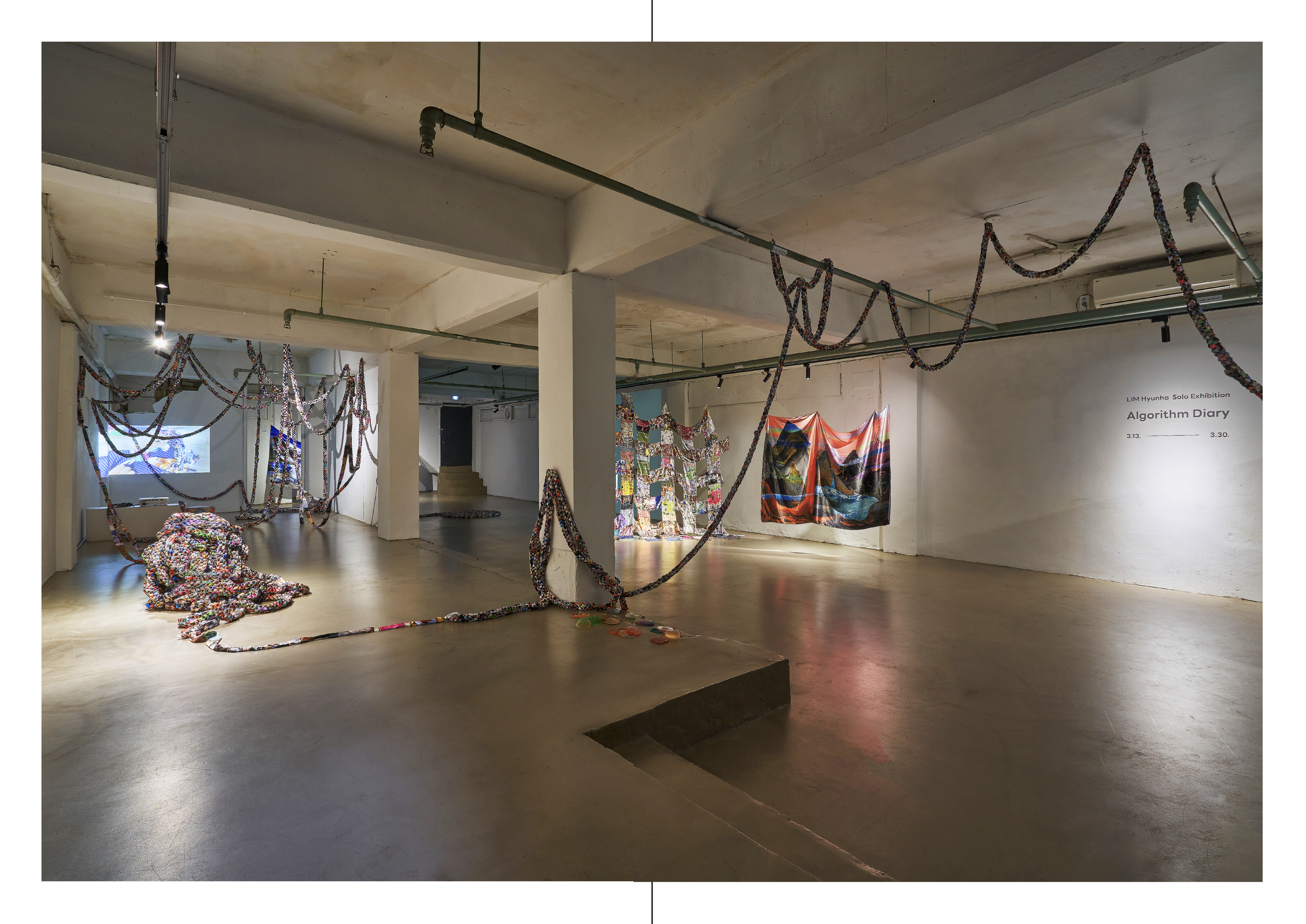 An art exhibition space with fabric and paper art installations hanging from the ceiling and displayed on the walls, including colorful woven and quilted pieces, with a sign indicating the exhibition title 'Algorithm Diary' by LIM Hyunha.