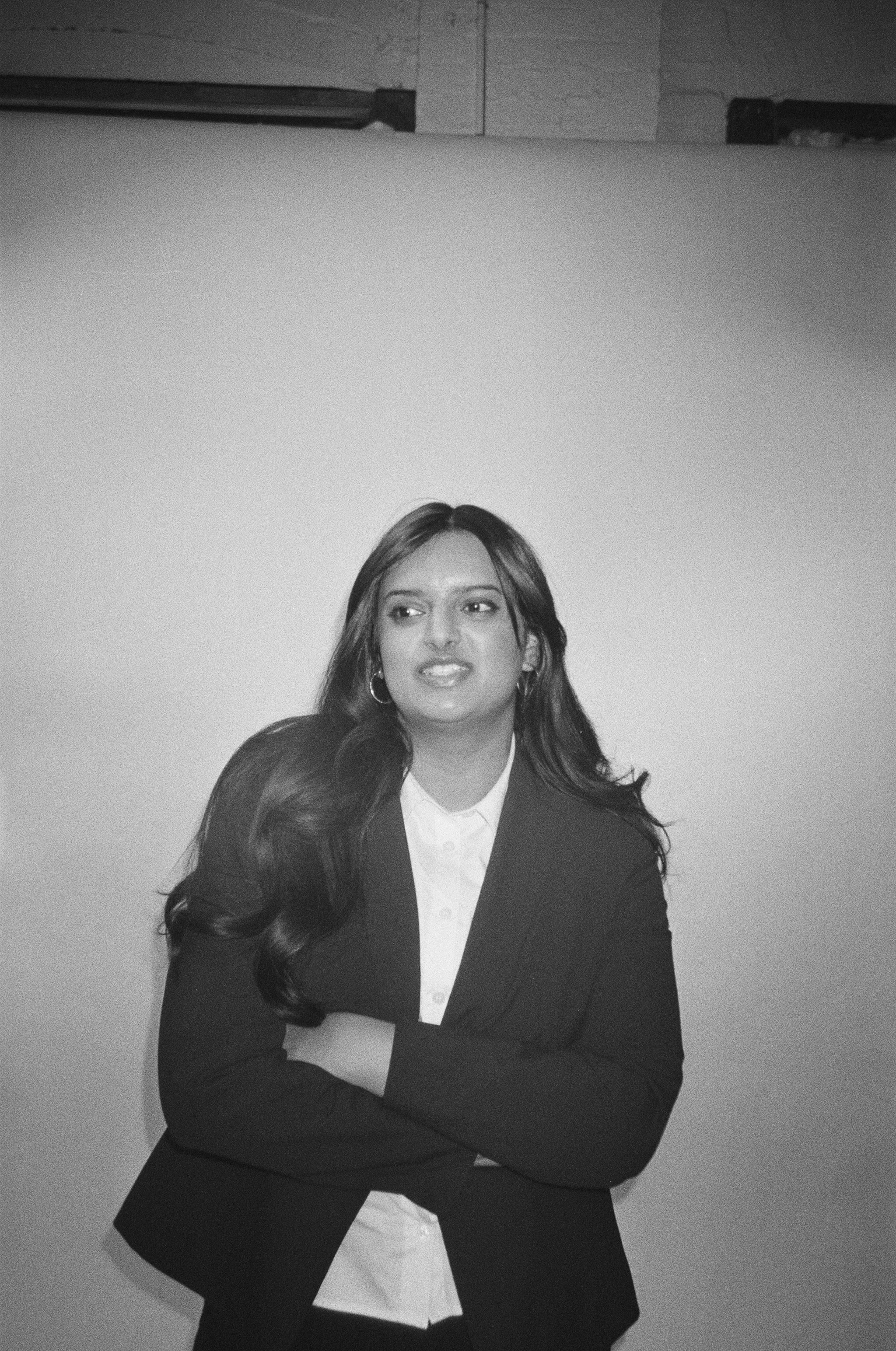 A woman with long, dark, wavy hair, wearing a white shirt and a dark blazer with arms crossed, standing against a plain wall with her head turned slightly to her left.