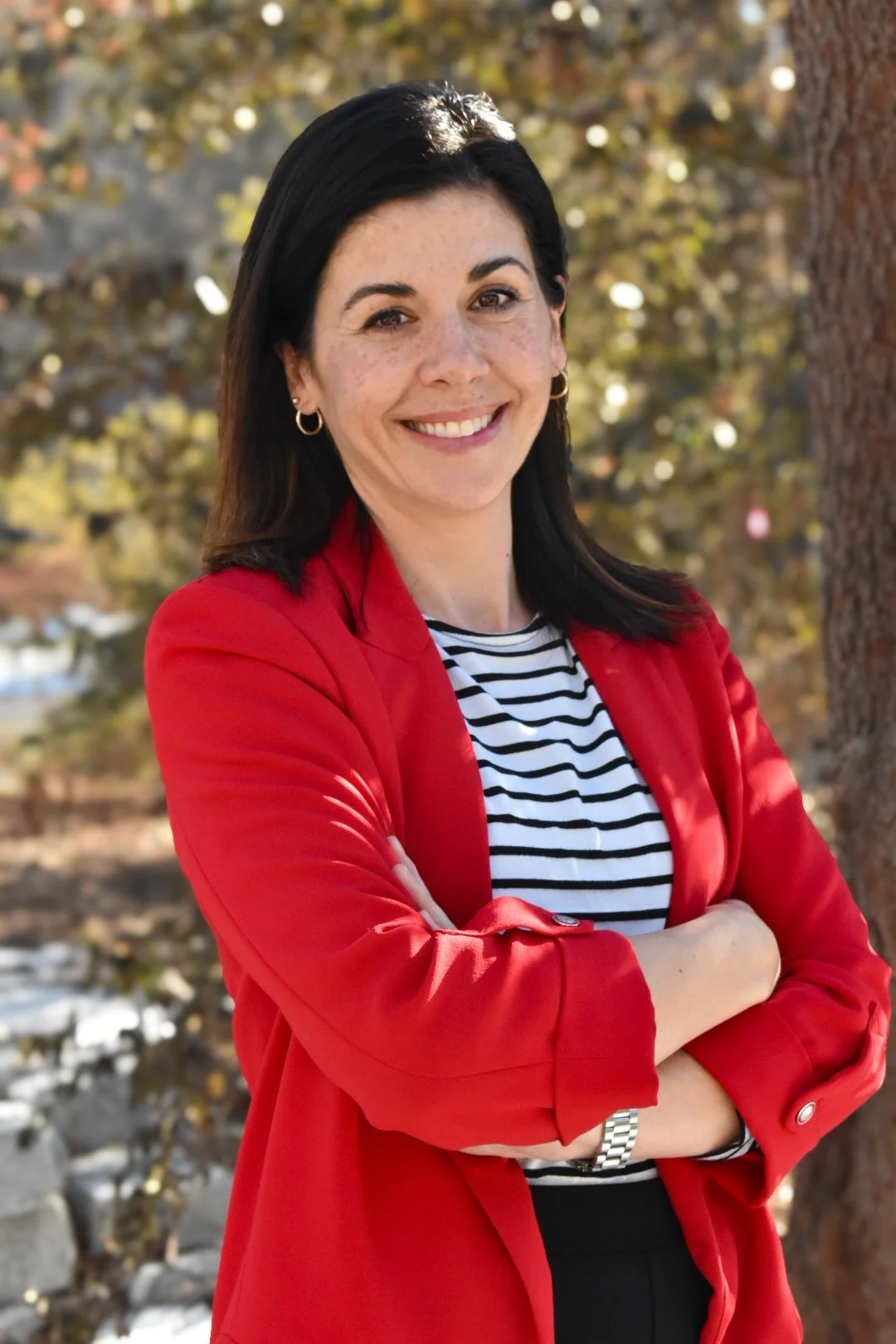 A woman with dark hair, wearing a red blazer, a black and white striped shirt, and a silver watch, standing outdoors with her arms crossed, smiling, with trees and sunlight in the background.