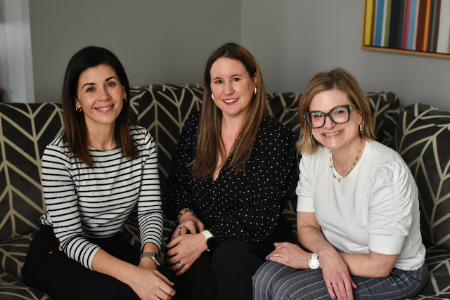 Three women sitting on a patterned black and beige sofa, smiling at the camera, in a room with a modern art piece on the wall.