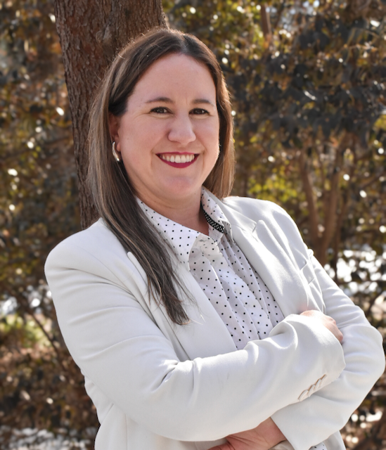 A woman with long brown hair and earrings, smiling with arms crossed, standing outdoors near a tree with autumn foliage, wearing a white blazer and a polka dot shirt.