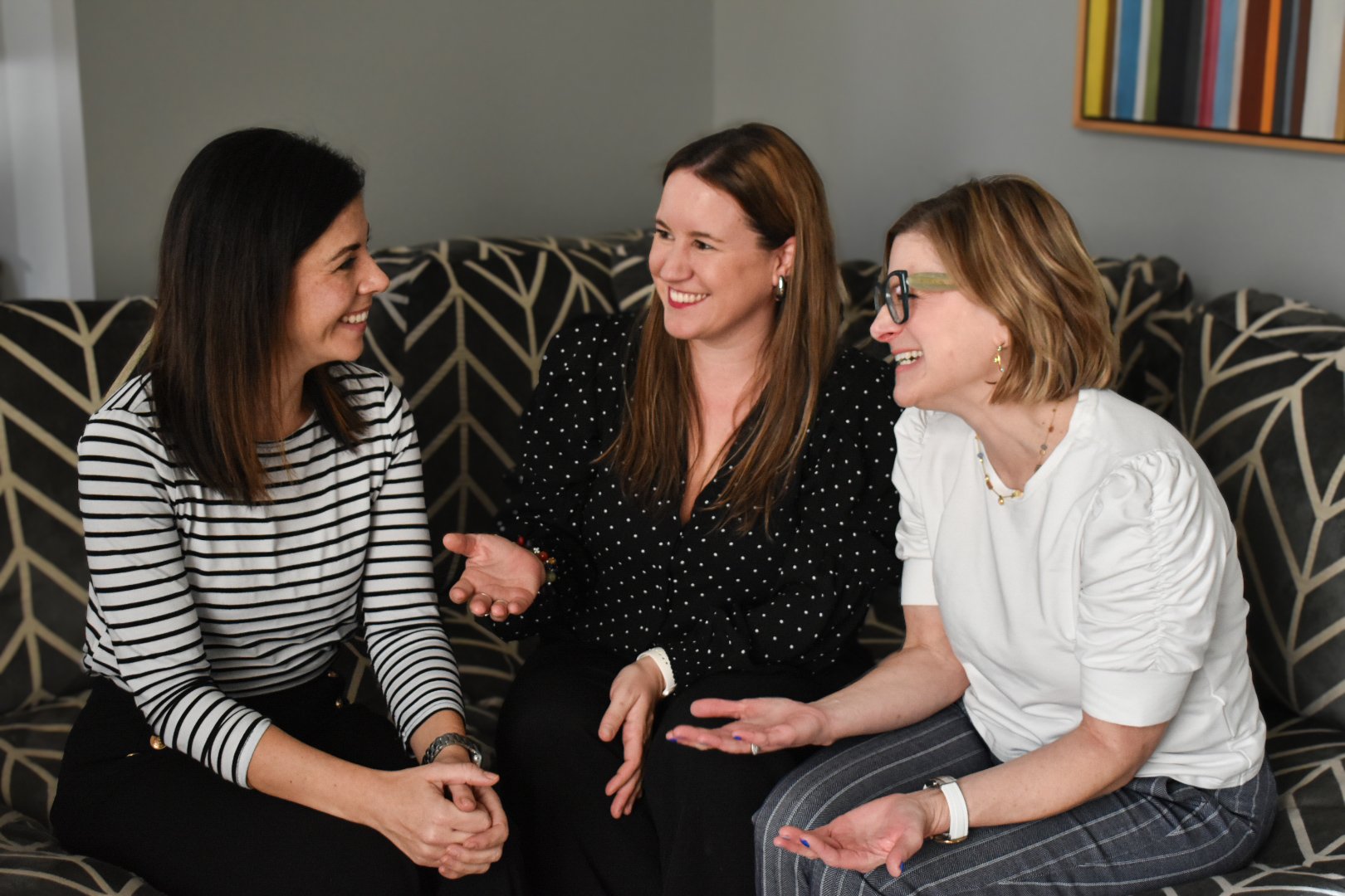 Three women sitting on a couch, engaging in a lively conversation and smiling happily.