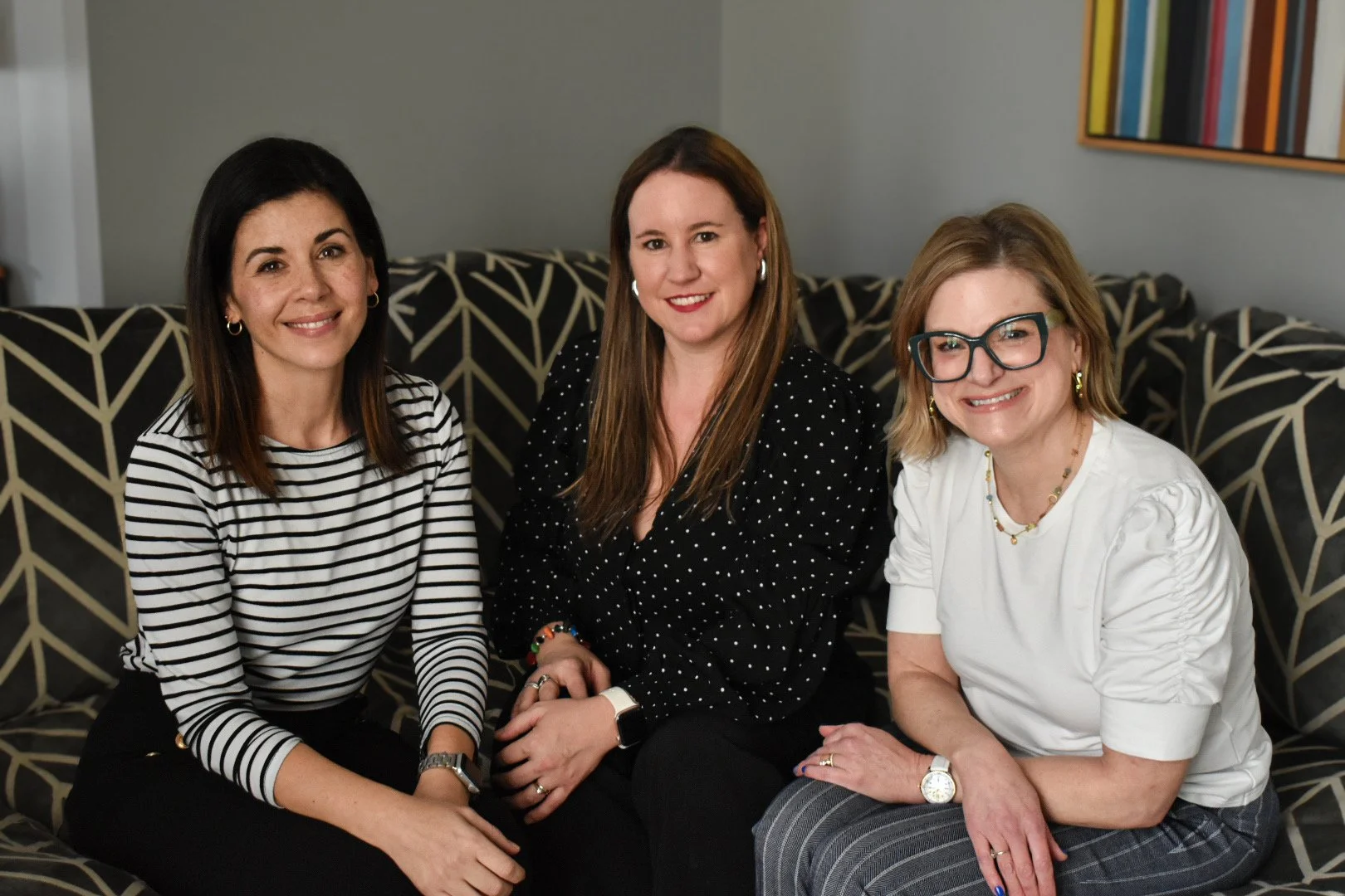 Three women sitting on a patterned couch, smiling at the camera in a living room with artwork on the wall.