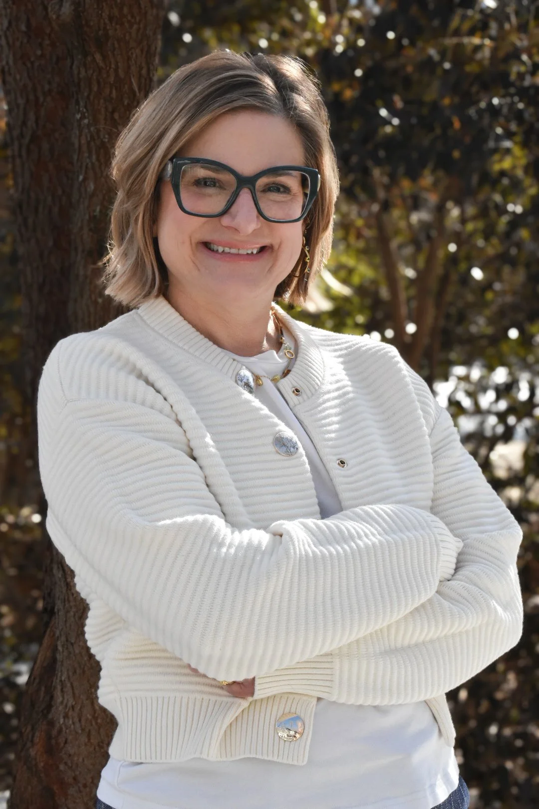 A woman with short blonde hair, wearing glasses, a white textured jacket, and a necklace, stands outdoors with trees and sunlight in the background.