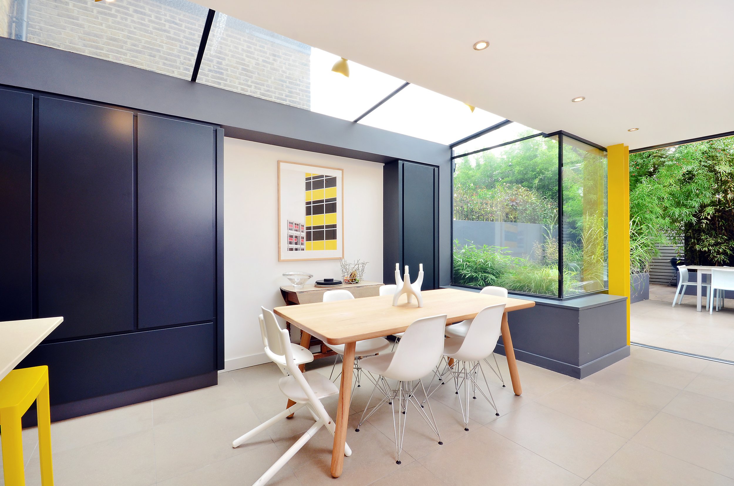 Modern dining room with a wooden table, white chairs, black wall cabinets, and large windows overlooking a green garden.