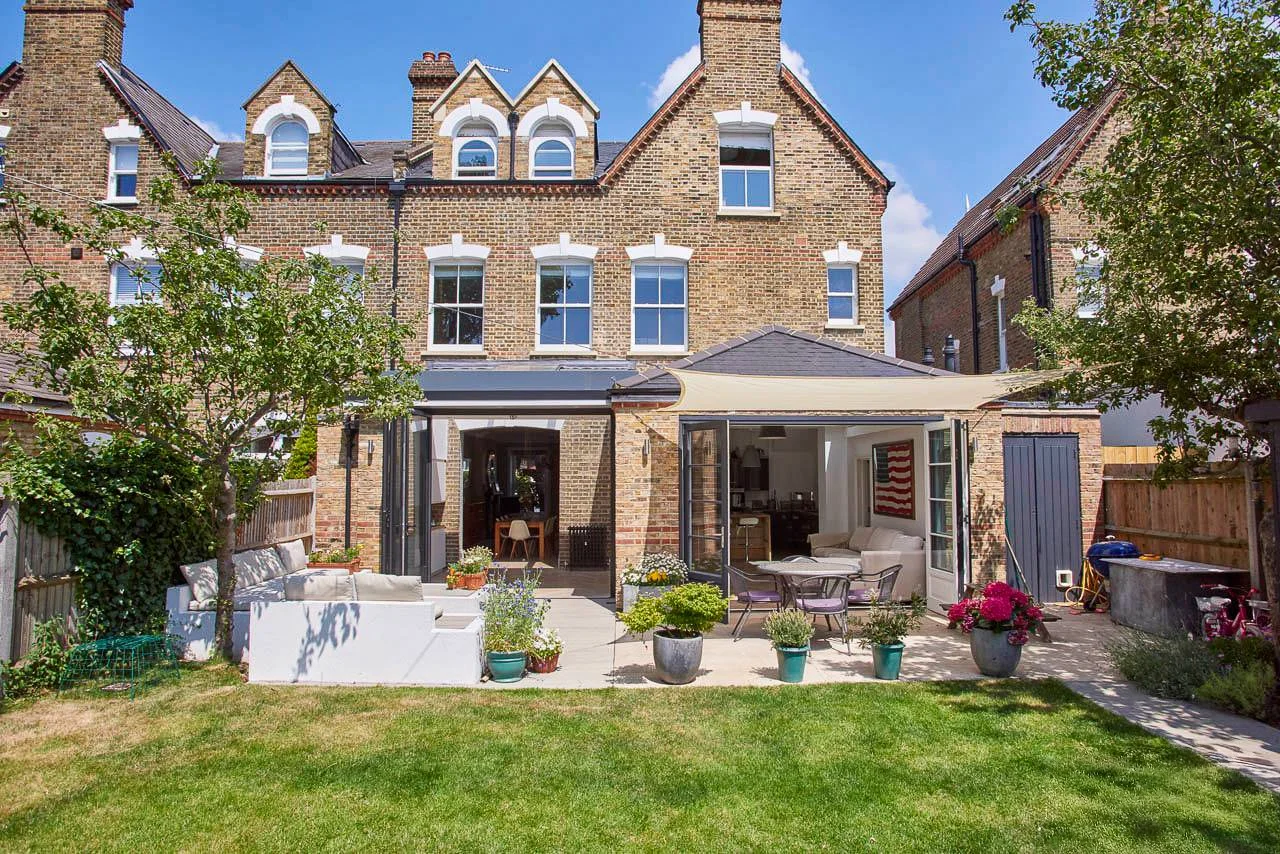 Backyard patio with outdoor furniture and potted plants, attached to a multi-story brick house with an extension and trees, under a partly cloudy sky.