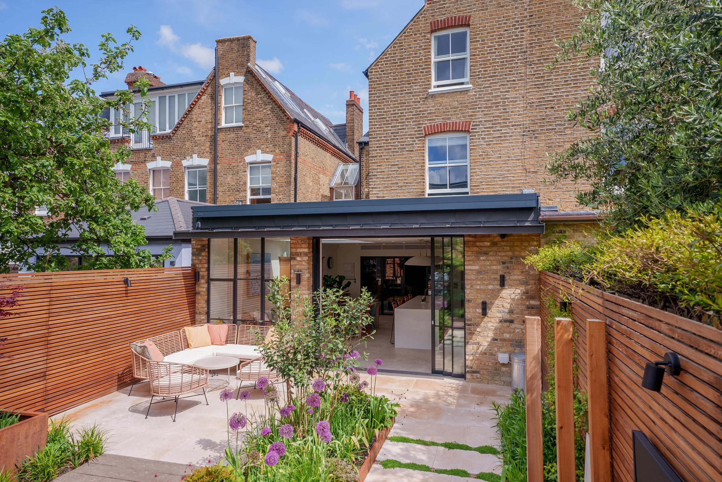 View of a modern backyard patio with outdoor seating, potted plants, and a glass sliding door leading to the interior of a brick house. The patio is enclosed by wooden fencing and surrounded by lush greenery. Residential buildings are visible in the background.