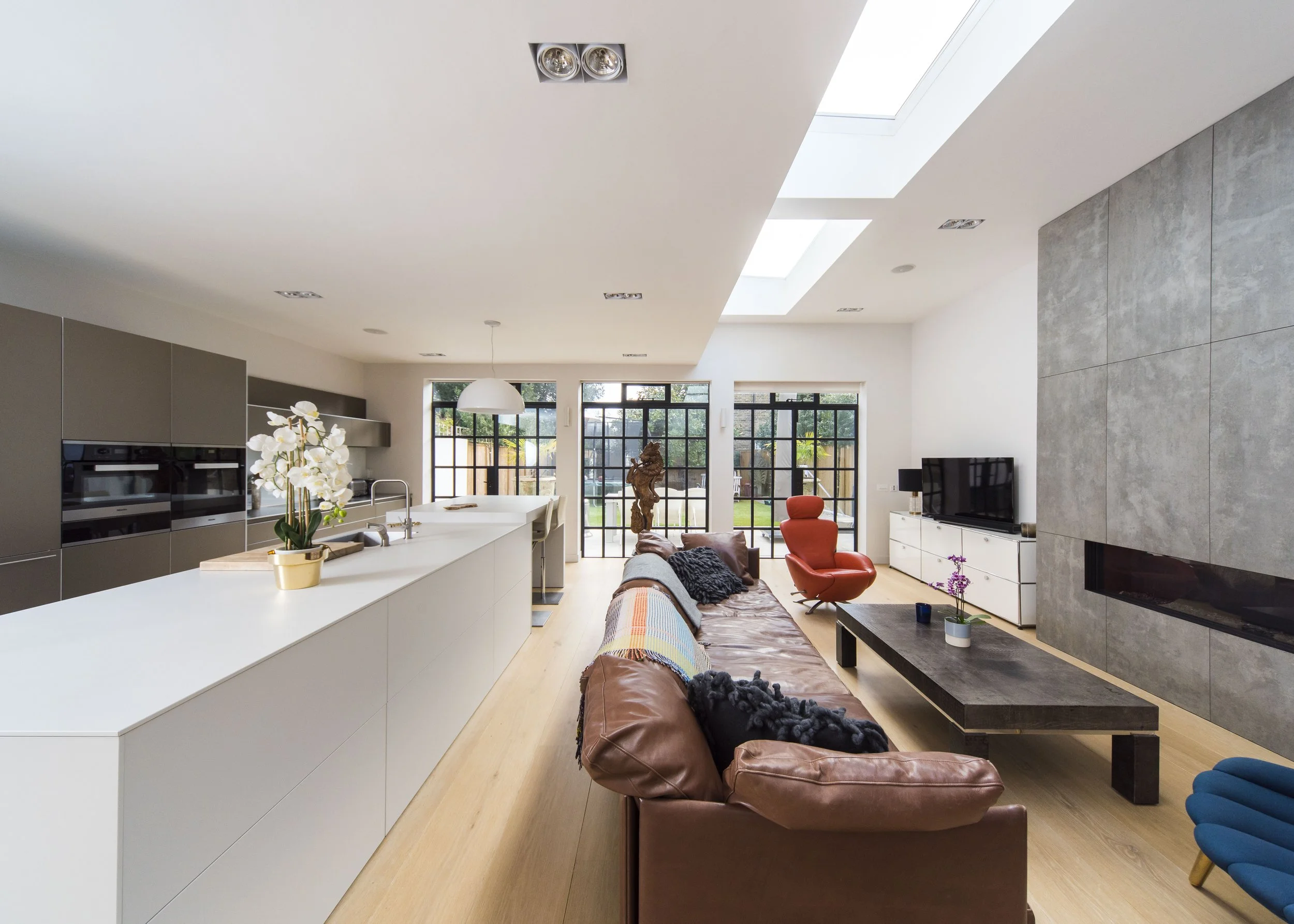 Modern open-plan living room and kitchen with natural light, brown leather sofa, colorful armchair, low wooden coffee table, white kitchen island, gray cabinetry, black window frames, and large skylights.