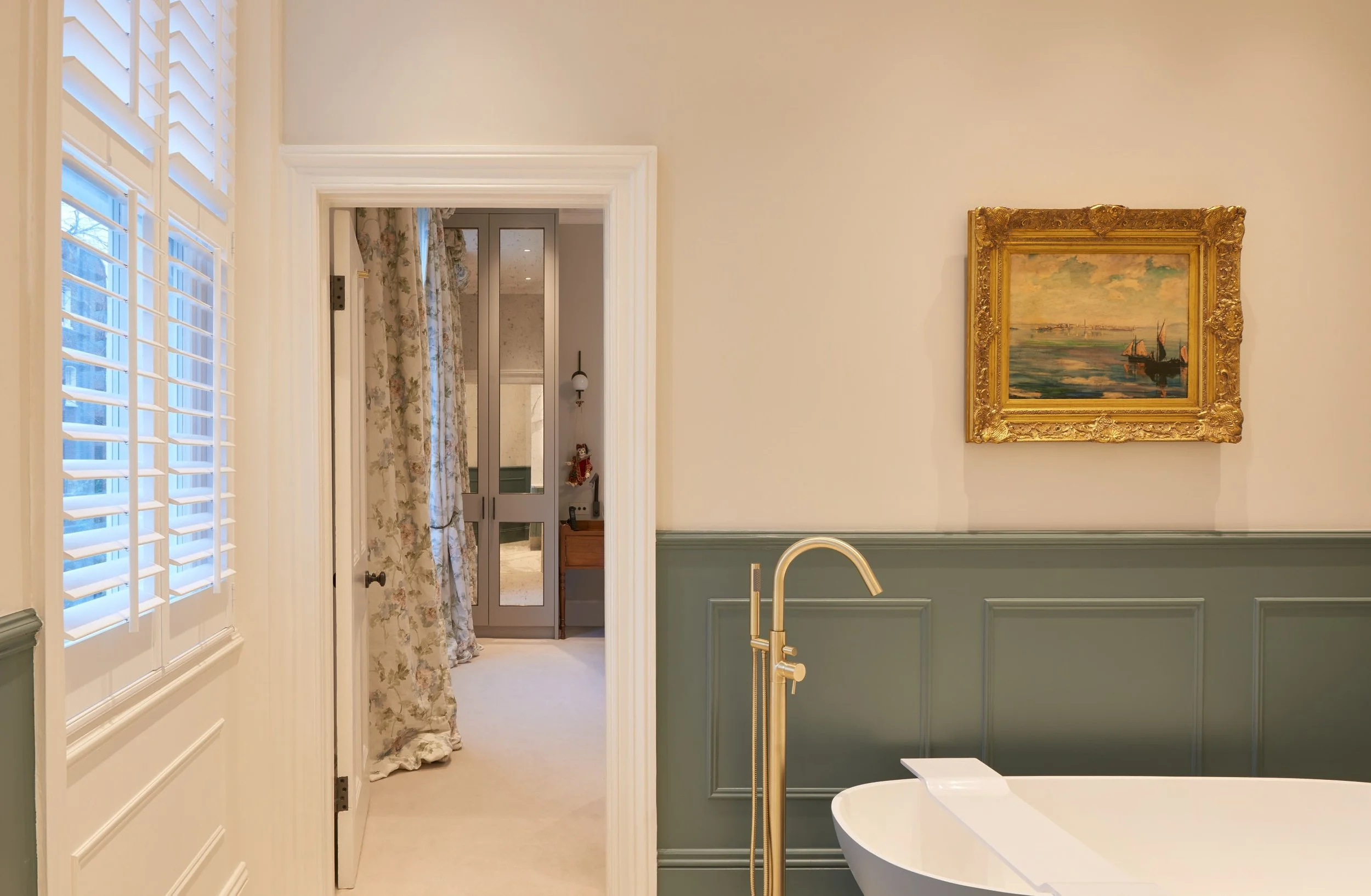 A bathroom with light green wainscoting, a white bathtub, and gold fixtures. A framed painting is hung on the white wall, and a window with white shutters is visible on the left. There is a doorway leading to a room with floral curtains and a gray door.