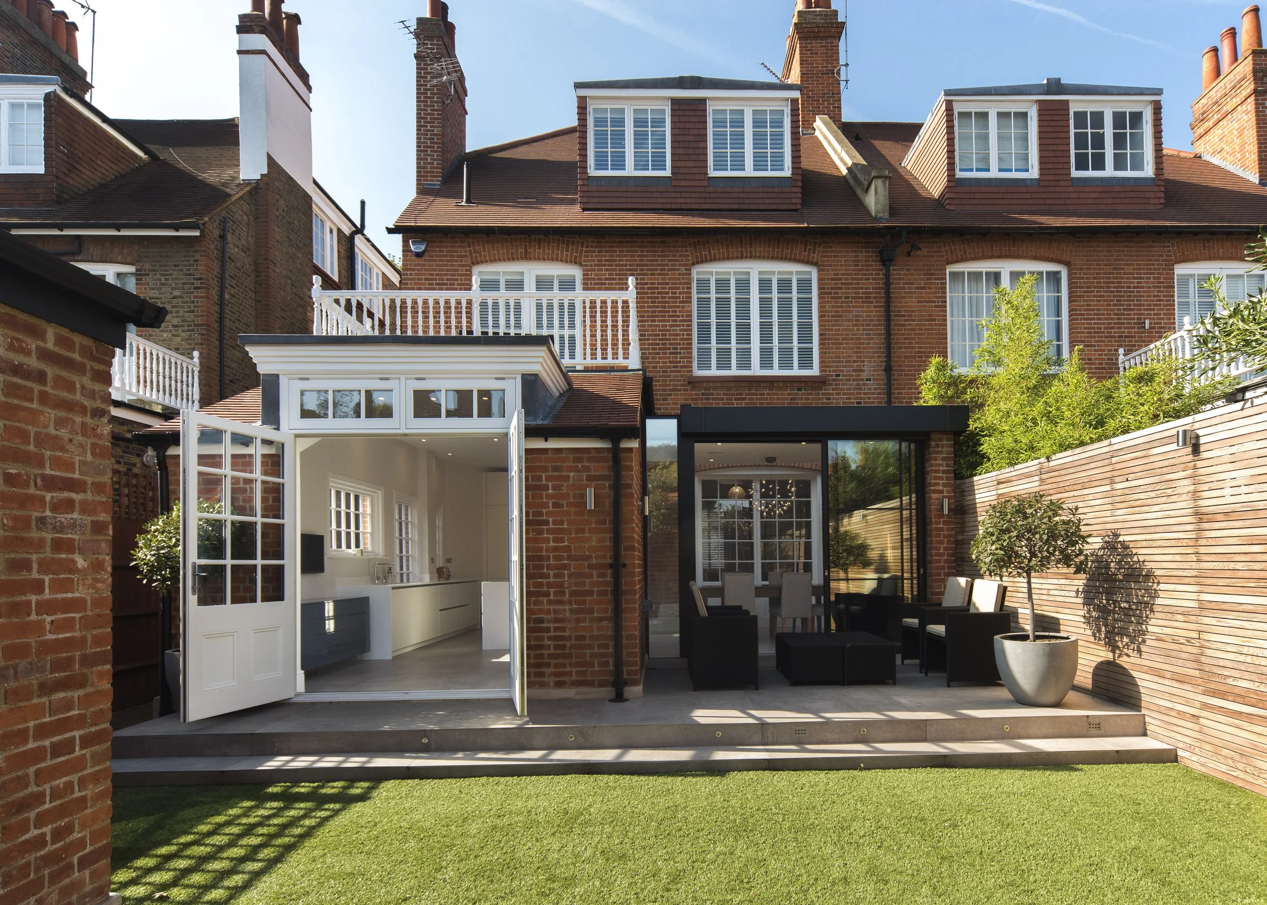 Backyard patio with outdoor seating and potted plants, connecting to interior kitchen and dining area through glass doors, with a brick wall and wooden fence on one side and neighboring brick houses in the background.