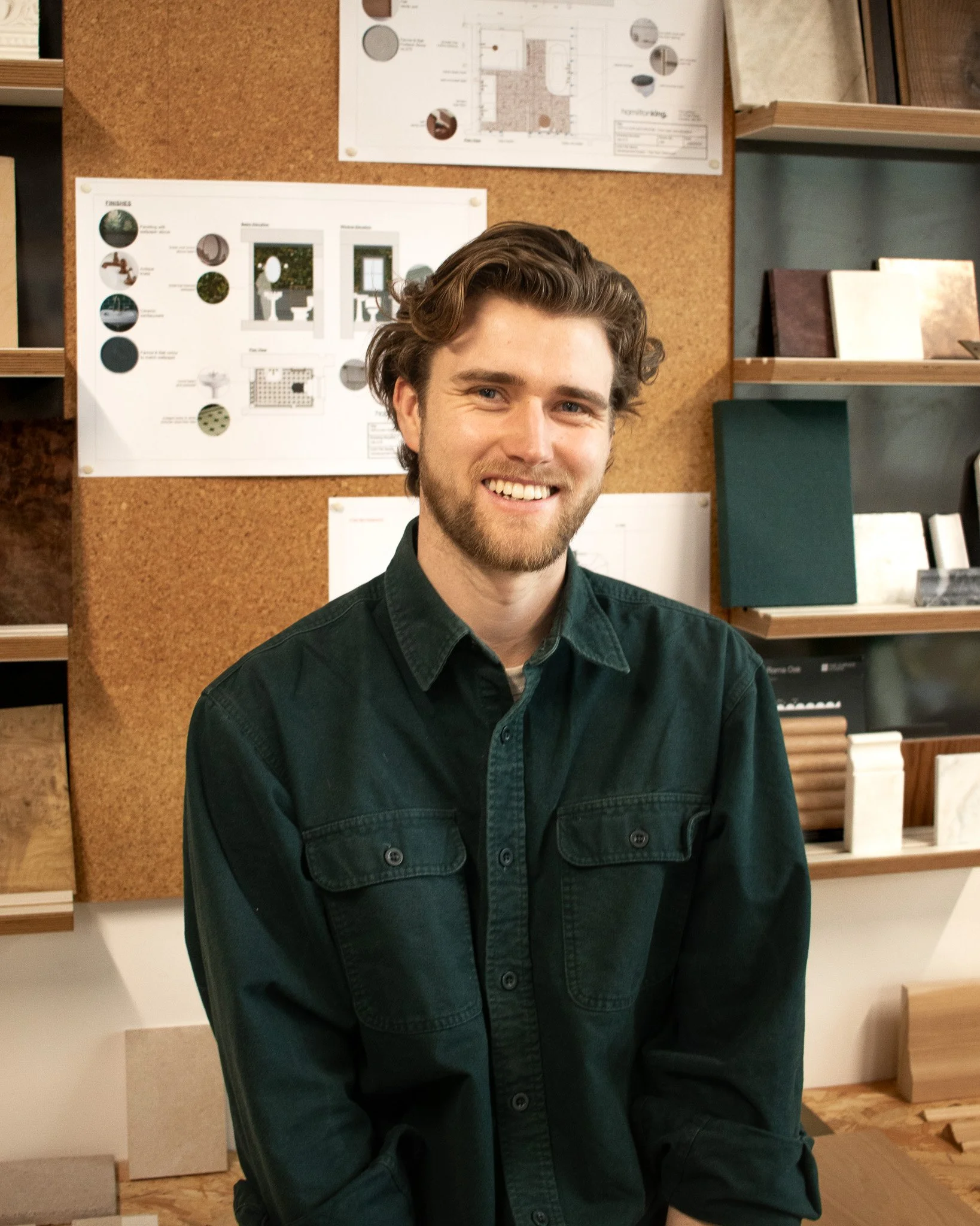 Tom has light skin, brown wavy hair, and a beard, smiling and sitting in a room with corkboard walls and architectural design plans. He is wearing a dark green button-up shirt.
