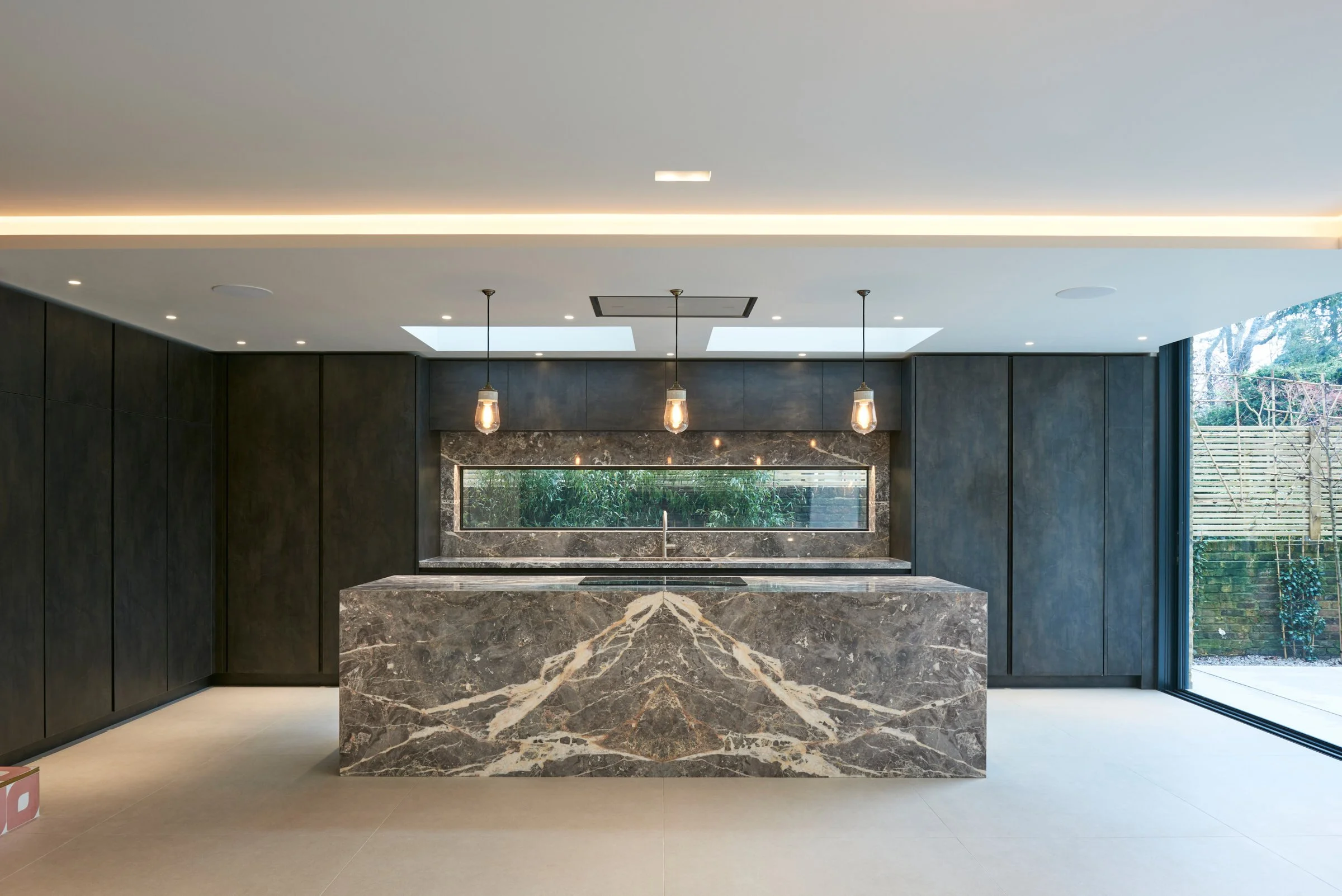 Modern kitchen with dark cabinetry, a marble island with beige, gray, and white veining, pendant lights, a long horizontal window with greenery outside, and large sliding glass door to the right leading to an outdoor area.