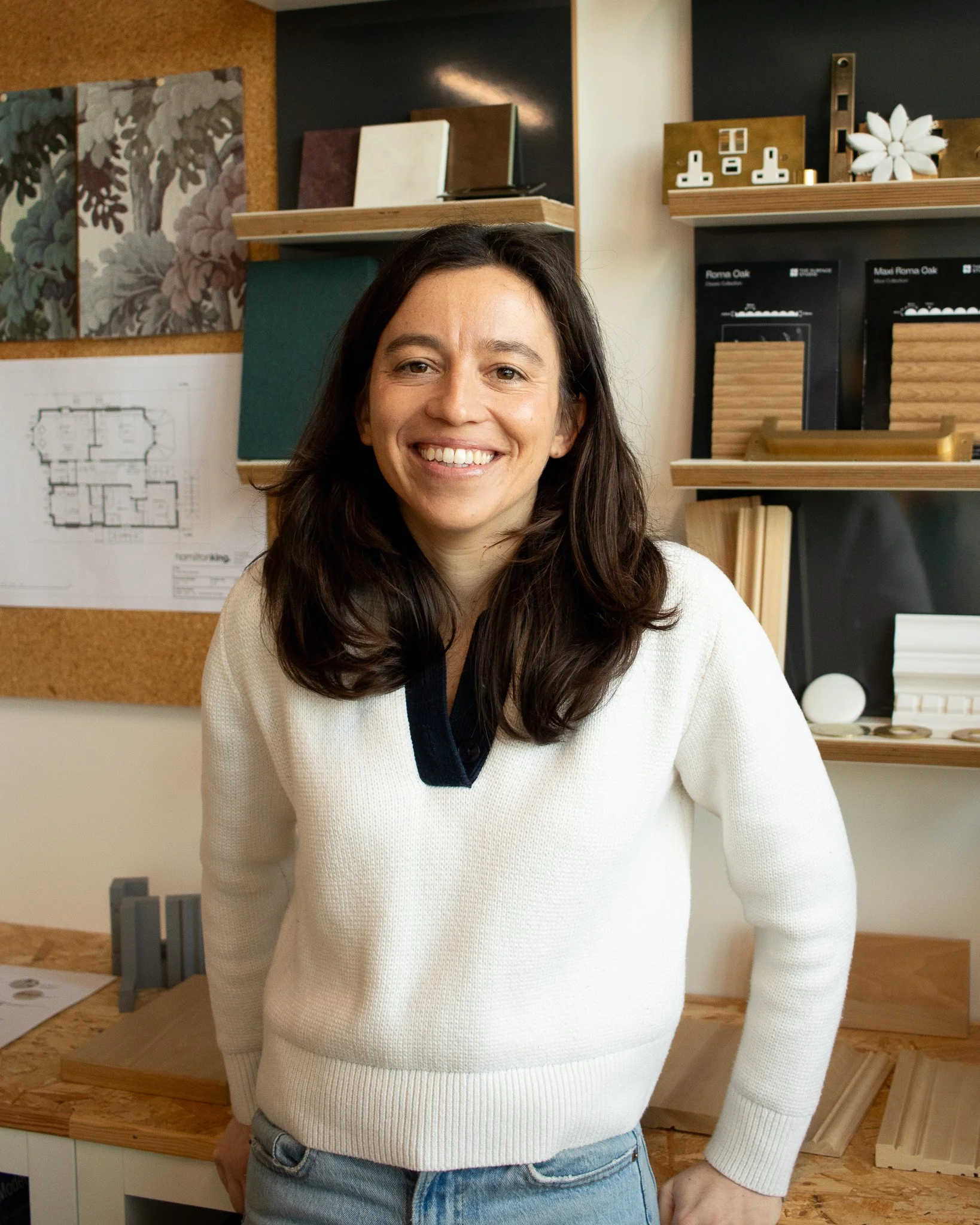 A woman with dark brown hair smiling in a architecture studio with design samples and architectural drawings in the background.