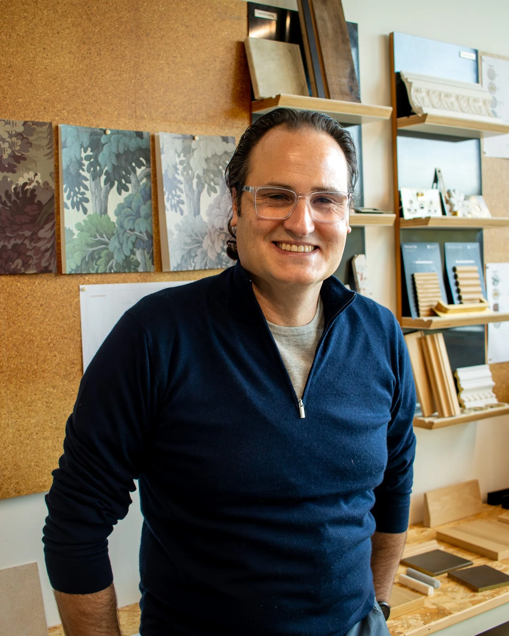 Jon has dark hair and glasses. He is smiling, standing in our design studio with shelves of samples and materials in the background.