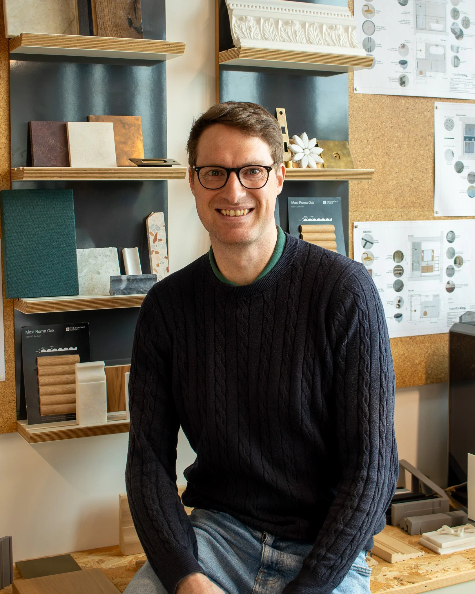 Tom is sitting on a wooden table in an interior & architecture studio, smiling at the camera, with shelves of color, material samples, and design plan posters on the wall behind him.