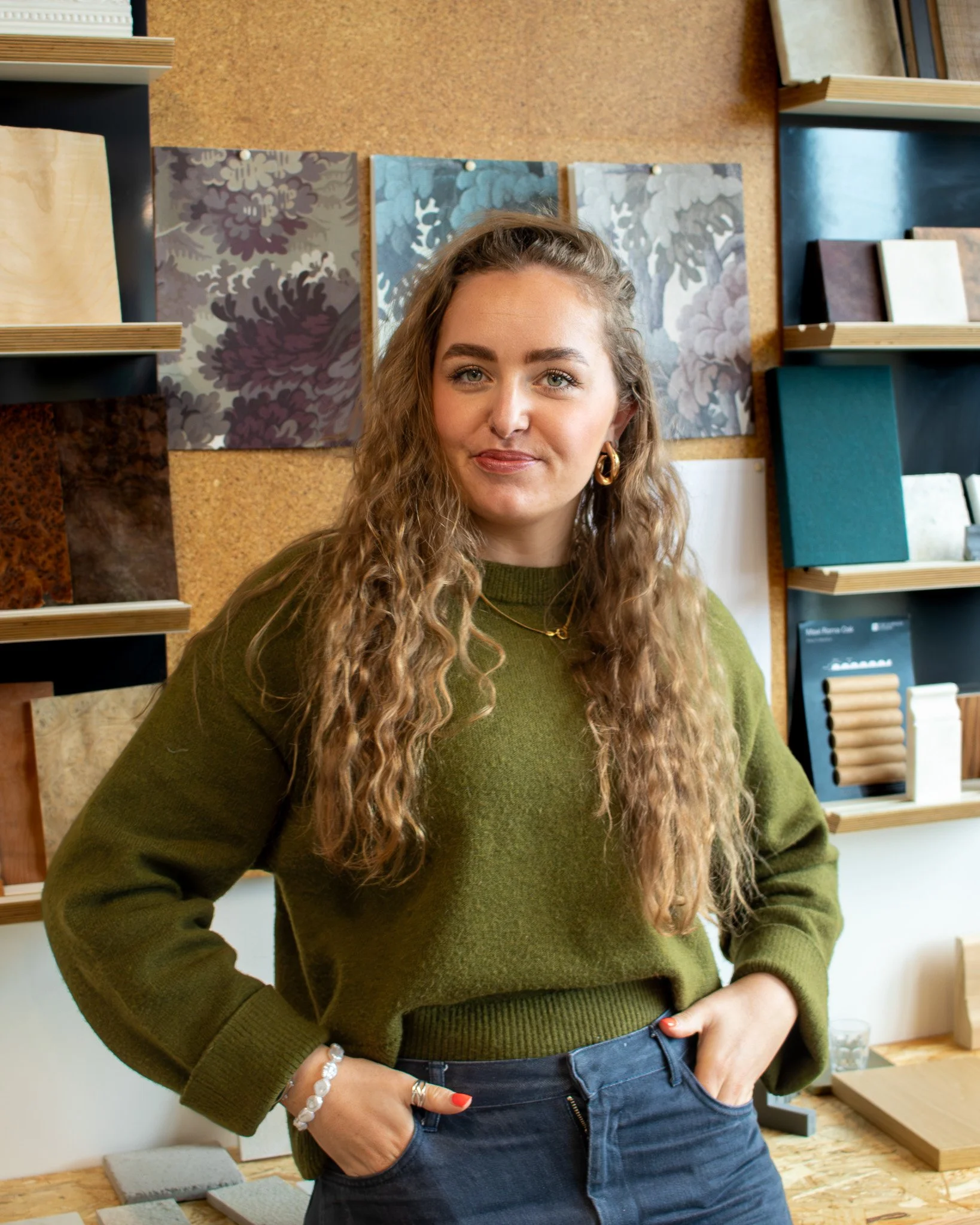 Hannah has long curly hair, & is wearing a green sweater and dark jeans, standing in front of a display with various sample materials and architectural drawings on a corkboard back wall.