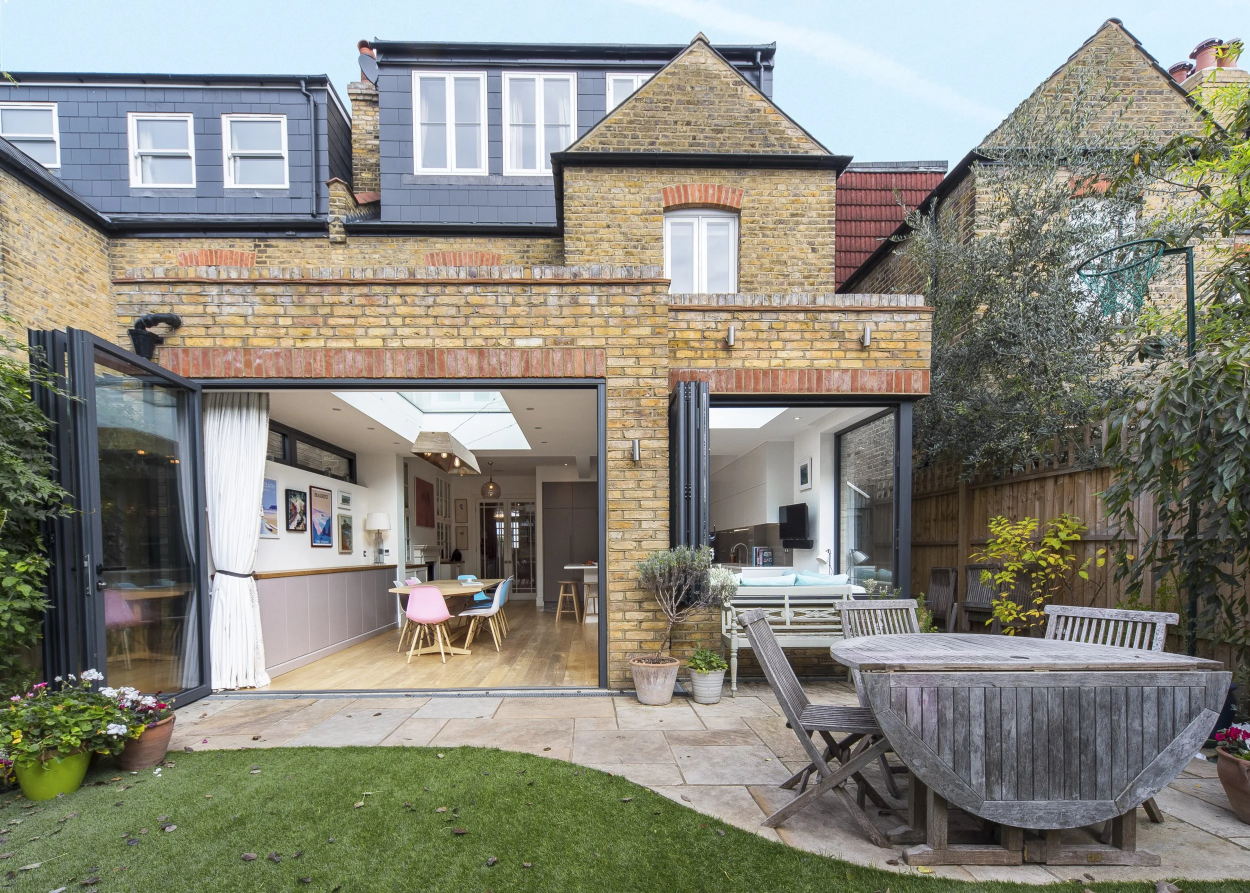 View of a brick house with large open sliding doors revealing a modern interior with dining and living areas, outdoor patio with wooden table and chairs, potted plants, and a lawn.
