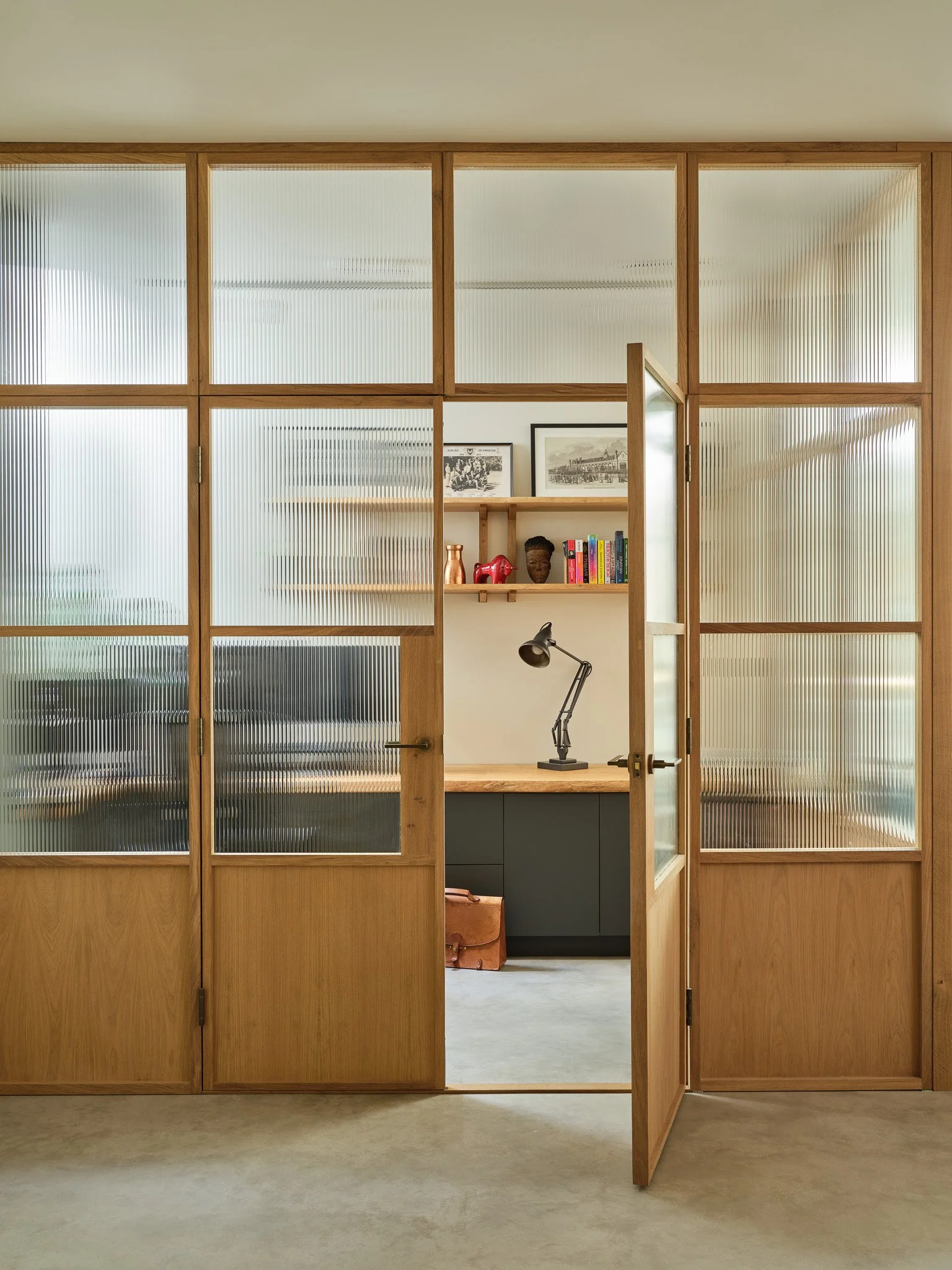 An office with a wooden double door, partially open, revealing a workspace with a desk lamp, books, and framed pictures on the wall.