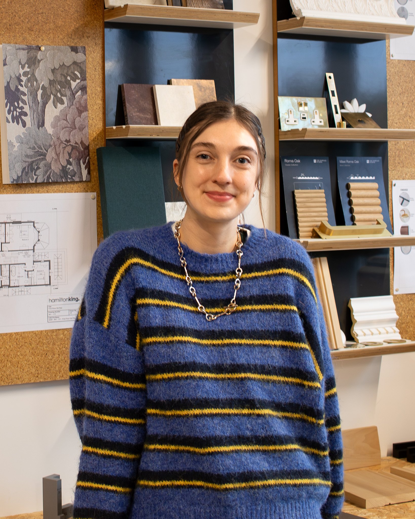 A young woman with brown hair in a braid, wearing a blue sweater with black and yellow stripes, and a silver chain necklace, standing indoors in front of a corkboard and shelves with architectural samples and drawings.