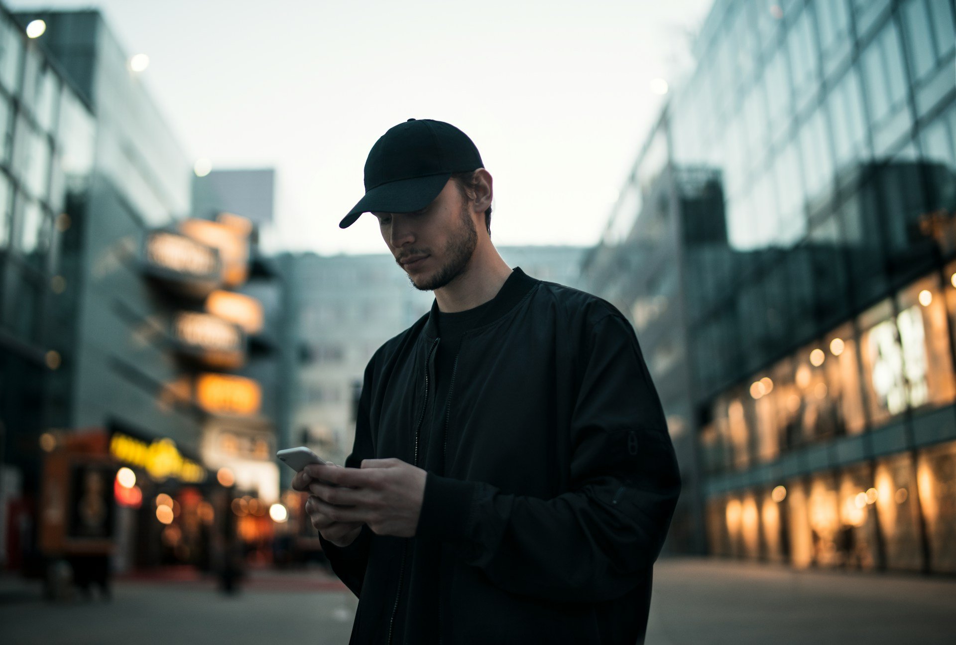 Man wearing black cap and jacket looking at cellphone in urban setting with modern glass buildings