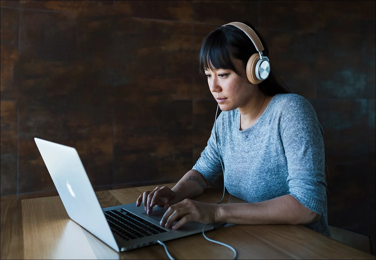 Woman wearing headphones working on a laptop at a wooden table.