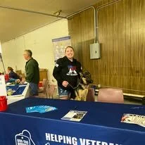 A smiling woman standing behind a table at a fundraising event for helping veterans, with a wooden wall in the background.