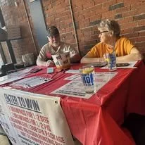Two young boys sitting at a table covered with a red tablecloth, eating and talking. One boy is wearing a white shirt with red stripes, and the other is wearing an orange shirt. There is a can of 7UP and some other items on the table.