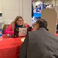 A woman with glasses and long hair sitting behind a table with a red tablecloth, facing a man in a gray jacket. They are indoors, possibly at a job fair or event.