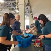 Four people working at a table, packing or preparing food items at a community event or food distribution.