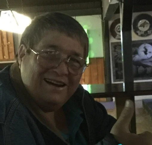 A man with glasses smiling in a dimly lit indoor space with framed pictures on the wall