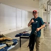A person wearing a baseball cap and a teal t-shirt standing in a room with shelves and a table, holding a black backpack. The room appears to be a classroom or storage space with clothing items on the table.
